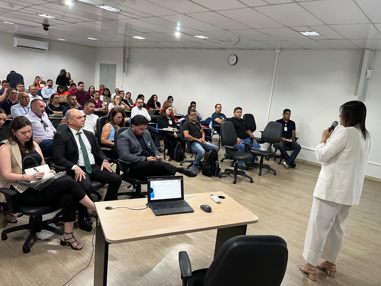 Imagem colorida mostra o interior de uma sala de aula com uma palestra em andamento. Uma mulher com cabelo escuro e vestindo um conjunto de blazer e calça brancos está de frente para o público, segurando um microfone na mão esquerda. A plateia, composta por homens e mulheres, está sentada em fileiras. Alguns participantes usam óculos, e a maioria está atenta à instrutora. No canto esquerdo da imagem, há uma mesa com um notebook aberto, um mouse e um controle. A sala é bem iluminada e tem um ar-condicionado na parede do fundo. Imagem colorida mostra o interior de uma sala de aula com uma palestra em andamento. Uma mulher com cabelo escuro e vestindo um conjunto de blazer e calça brancos está de frente para o público, segurando um microfone na mão esquerda. A plateia, composta por homens e mulheres, está sentada em fileiras. Alguns participantes usam óculos, e a maioria está atenta à instrutora. No canto esquerdo da imagem, há uma mesa com um notebook aberto, um mouse e um controle. A sala é bem iluminada e tem um ar-condicionado na parede do fundo.