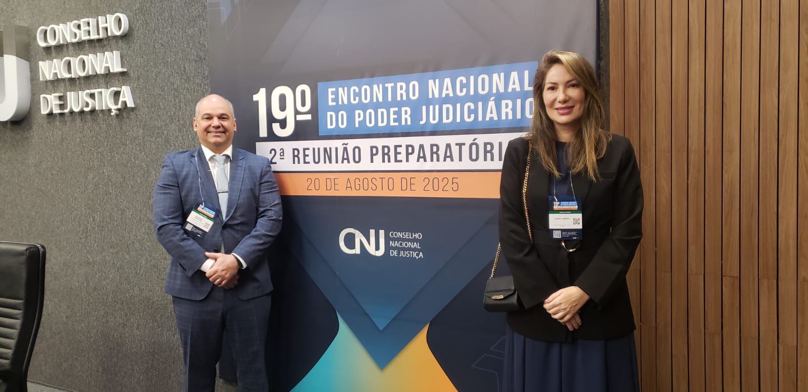 Imagem colorida mostra duas pessoas, um homem e uma mulher, em pé em frente a um banner. O homem, careca e de terno azul-marinho, usa um crachá no peito. A mulher, com cabelo castanho solto, usa um blazer preto e uma saia longa azul. O banner tem um logo que diz "Conselho Nacional de Justiça" e o título "19º Encontro Nacional do Poder Judiciário, 2ª Reunião Preparatória, 20 de agosto de 2025". Imagem colorida mostra duas pessoas, um homem e uma mulher, em pé em frente a um banner. O homem, careca e de terno azul-marinho, usa um crachá no peito. A mulher, com cabelo castanho solto, usa um blazer preto e uma saia longa azul. O banner tem um logo que diz "Conselho Nacional de Justiça" e o título "19º Encontro Nacional do Poder Judiciário, 2ª Reunião Preparatória, 20 de agosto de 2025".