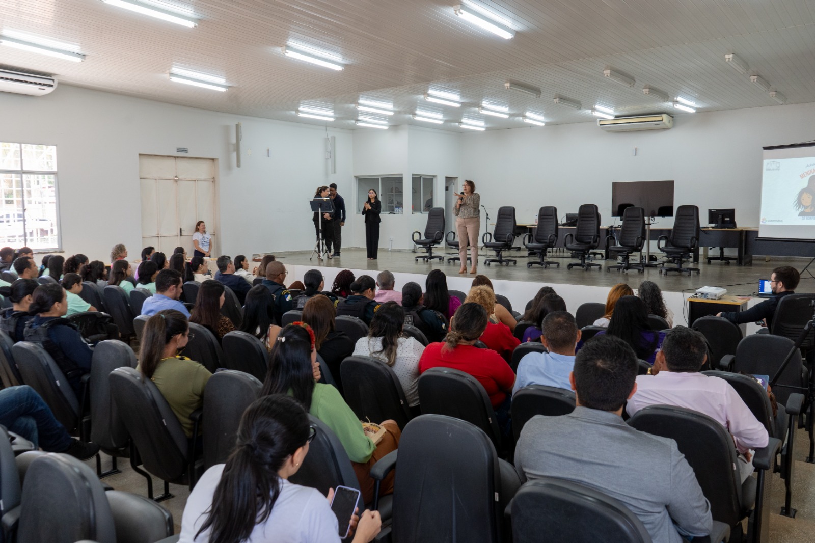 fotografia tirada de frente, mostra um grande auditório com um público em cadeiras pretas assistindo a uma palestra. No palco, mais elevado, há um painel de cadeiras vazias à direita e, ao centro, três pessoas em pé, sendo uma delas uma mulher falando a um microfone.