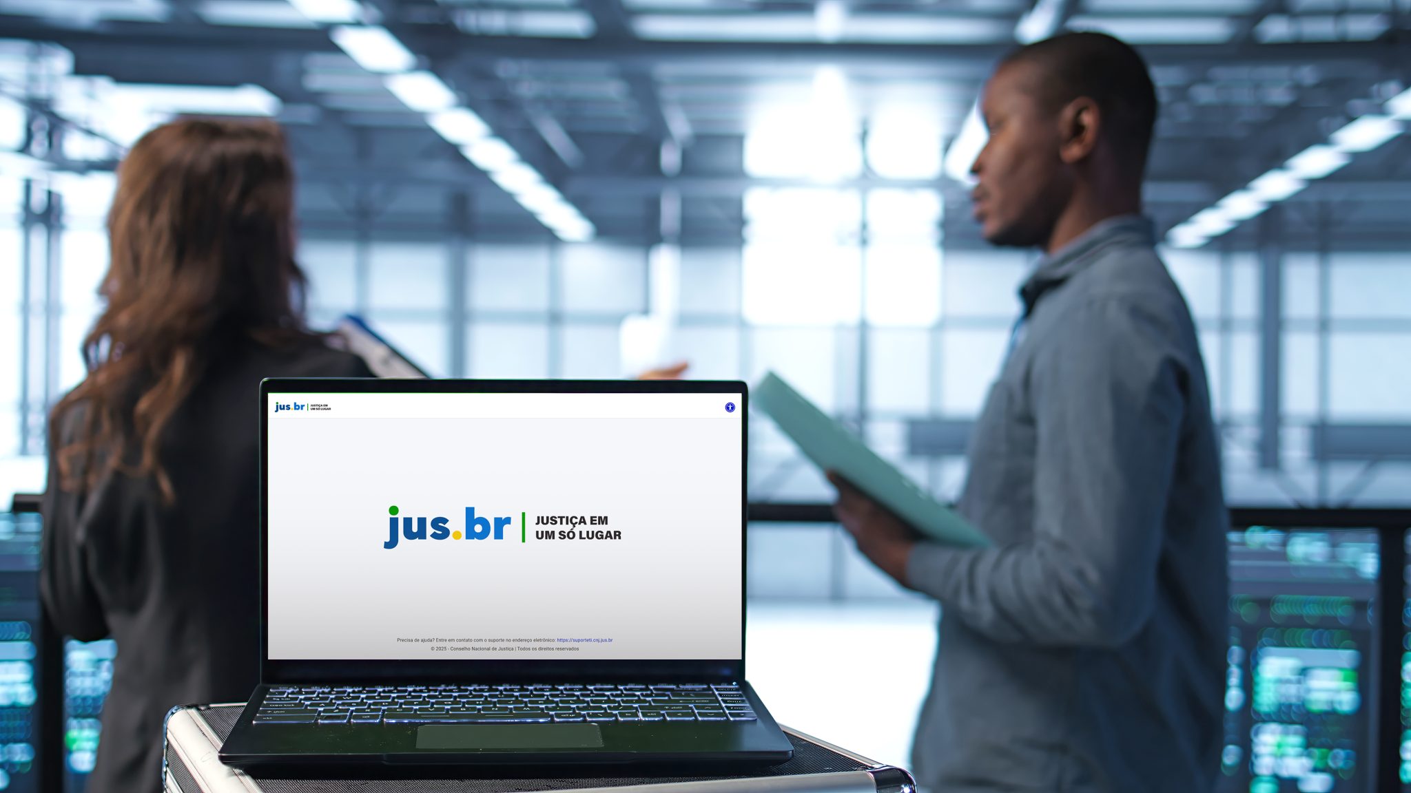 Foto colorida mostra em primeiro plano, um notebook aberto exibindo a página inicial do portal jus.br, com o logotipo do site no centro da tela e o slogan “Justiça em um só lugar” ao lado direito. O plano de fundo é um ambiente corporativo moderno, com iluminação natural e painéis de dados ao fundo. Duas pessoas estão em segundo plano, desfocadas: uma mulher de cabelos longos, vista de costas, e um homem segurando uma pasta verde. Foto colorida mostra em primeiro plano, um notebook aberto exibindo a página inicial do portal jus.br, com o logotipo do site no centro da tela e o slogan “Justiça em um só lugar” ao lado direito. O plano de fundo é um ambiente corporativo moderno, com iluminação natural e painéis de dados ao fundo. Duas pessoas estão em segundo plano, desfocadas: uma mulher de cabelos longos, vista de costas, e um homem segurando uma pasta verde.