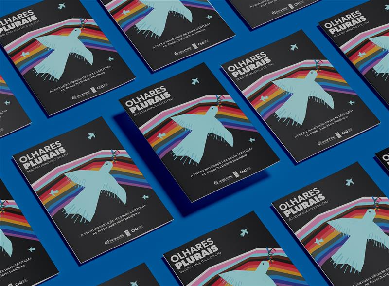 Diversos exemplares de uma mesma publicação impressa estão dispostos em padrão repetido sobre uma superfície azul. A capa dos materiais traz o título “OLHARES PLURAIS – Boletim Analítico do CNJ” em letras brancas sobre fundo preto. Ao centro, há a ilustração de uma pomba azul clara em voo, com o olho em destaque, posicionada sobre uma faixa diagonal formada pelas cores da bandeira LGBTQIA+. No canto inferior da capa, aparece o texto “A institucionalização da pauta LGBTQIA+ no Poder Judiciário brasileiro”. Abaixo do título, estão os logotipos do Conselho Nacional de Justiça (CNJ) e do Observatório de Direitos Humanos do Poder Judiciário.