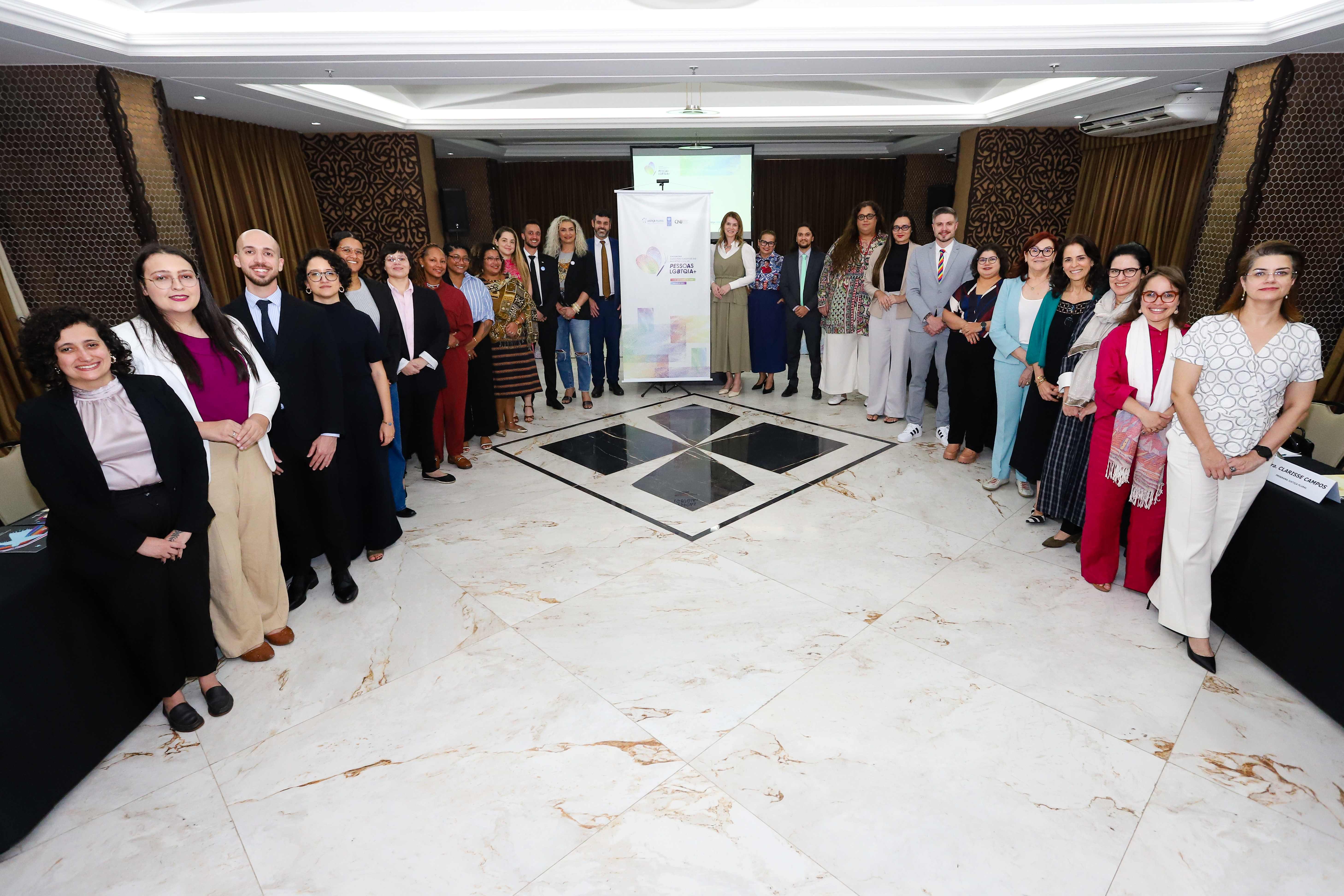 Foto mostra um grupo diverso de pessoas posando para a foto em uma sala ampla e bem iluminada, com piso de mármore claro e cortinas escuras ao fundo. Os participantes estão dispostos em duas fileiras laterais, formando um corredor central que leva a um banner institucional posicionado ao fundo. Durante sua participação, a magistrada ressaltou o mutirão “Trans‑Formando Identidades”, iniciativa da Justiça Itinerante em parceria com a Defensoria Pública de Roraima (DPE-RR), que busca facilitar o acesso ao registro civil e garantir direitos fundamentais dessa população. Foto mostra um grupo diverso de pessoas posando para a foto em uma sala ampla e bem iluminada, com piso de mármore claro e cortinas escuras ao fundo. Os participantes estão dispostos em duas fileiras laterais, formando um corredor central que leva a um banner institucional posicionado ao fundo. Durante sua participação, a magistrada ressaltou o mutirão “Trans‑Formando Identidades”, iniciativa da Justiça Itinerante em parceria com a Defensoria Pública de Roraima (DPE-RR), que busca facilitar o acesso ao registro civil e garantir direitos fundamentais dessa população.