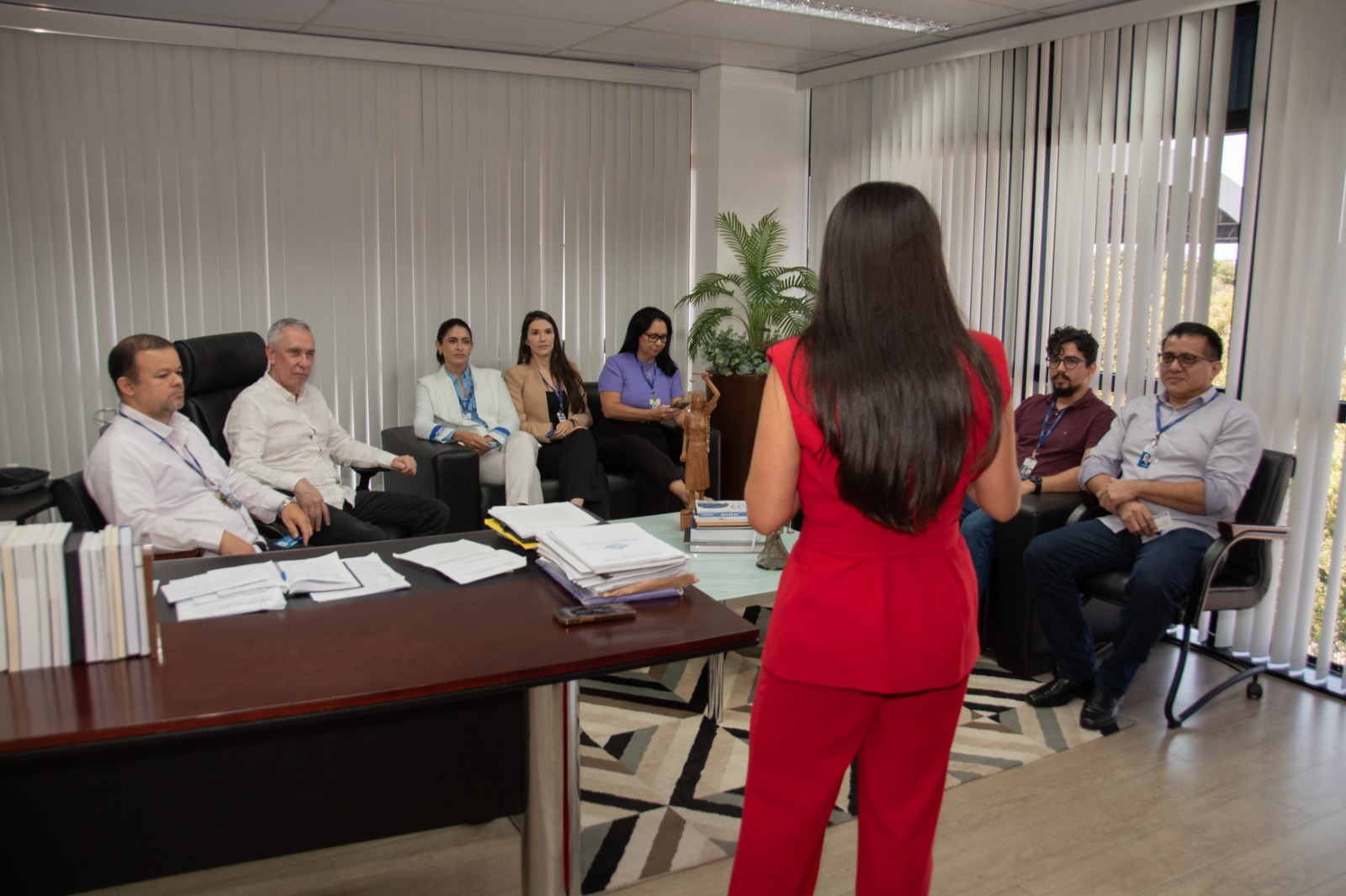 Foto colorida mostra uma sala de gabinete com oito pessoas reunidas. Em primeiro plano, uma mulher de cabelos longos, lisos e escuros, veste conjunto social vermelho e está de costas, falando para o grupo. À esquerda, há uma mesa de madeira com pilhas de documentos e livros. Em frente à mulher, estão sete pessoas sentadas em cadeiras e poltronas: quatro homens e três mulheres, todas vestidas com roupas formais.