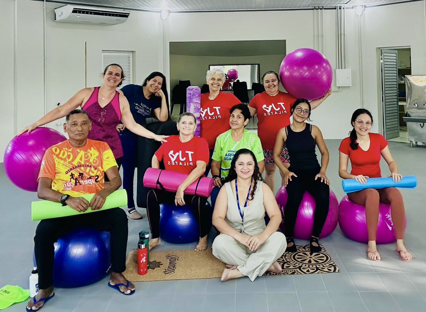 Imagem colorida mostra um grupo de dez pessoas em uma sala de ginástica. Elas estão sentadas ou de pé, cercadas por grandes bolas de Pilates e tapetes de ioga. Um homem está sentado à esquerda em uma bola com um tapete no colo. No centro, algumas mulheres estão de pé e uma está sentada entre elas. À direita, outras mulheres estão sentadas em bolas de Pilates, segurando tapetes de ioga. Outra mulher está sentada no chão entre os dois grupos. Todos parecem relaxados e felizes, posando para a foto em um ambiente de luz clara. Imagem colorida mostra um grupo de dez pessoas em uma sala de ginástica. Elas estão sentadas ou de pé, cercadas por grandes bolas de Pilates e tapetes de ioga. Um homem está sentado à esquerda em uma bola com um tapete no colo. No centro, algumas mulheres estão de pé e uma está sentada entre elas. À direita, outras mulheres estão sentadas em bolas de Pilates, segurando tapetes de ioga. Outra mulher está sentada no chão entre os dois grupos. Todos parecem relaxados e felizes, posando para a foto em um ambiente de luz clara.
