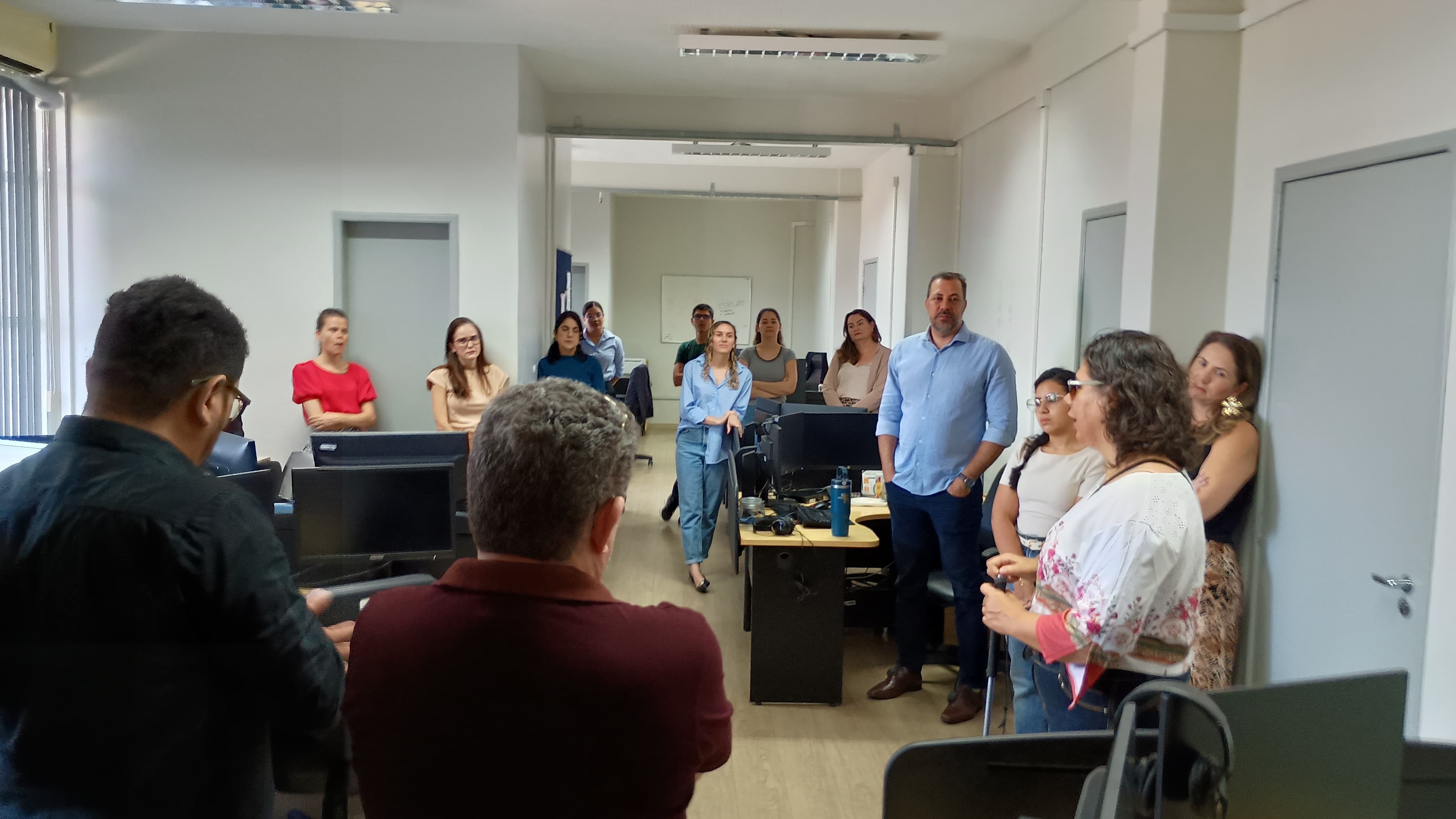 Imagem colorida mostra um escritório. Algumas pessoas estão sentadas em mesas com computadores, enquanto outras estão em pé, observando. O ambiente é um escritório de trabalho com divisórias e portas ao fundo. Imagem colorida mostra um escritório. Algumas pessoas estão sentadas em mesas com computadores, enquanto outras estão em pé, observando. O ambiente é um escritório de trabalho com divisórias e portas ao fundo.