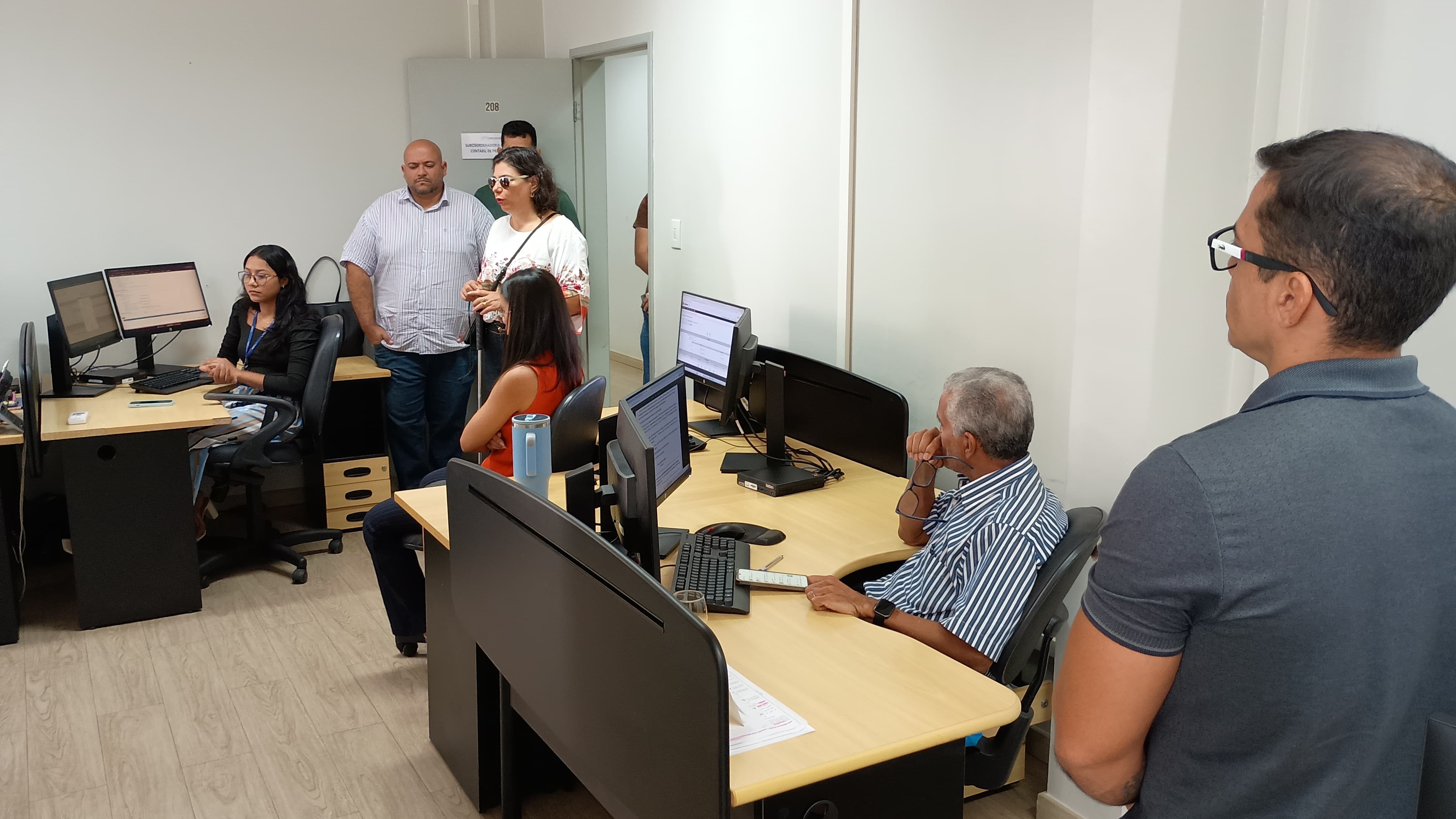 Imagem colorida mostra um escritório onde várias pessoas estão reunidas em pé, olhando para a frente. O ambiente parece ser um escritório com mesas e computadores ao fundo. A maioria das pessoas está vestindo roupas casuais. Imagem colorida mostra um escritório onde várias pessoas estão reunidas em pé, olhando para a frente. O ambiente parece ser um escritório com mesas e computadores ao fundo. A maioria das pessoas está vestindo roupas casuais.