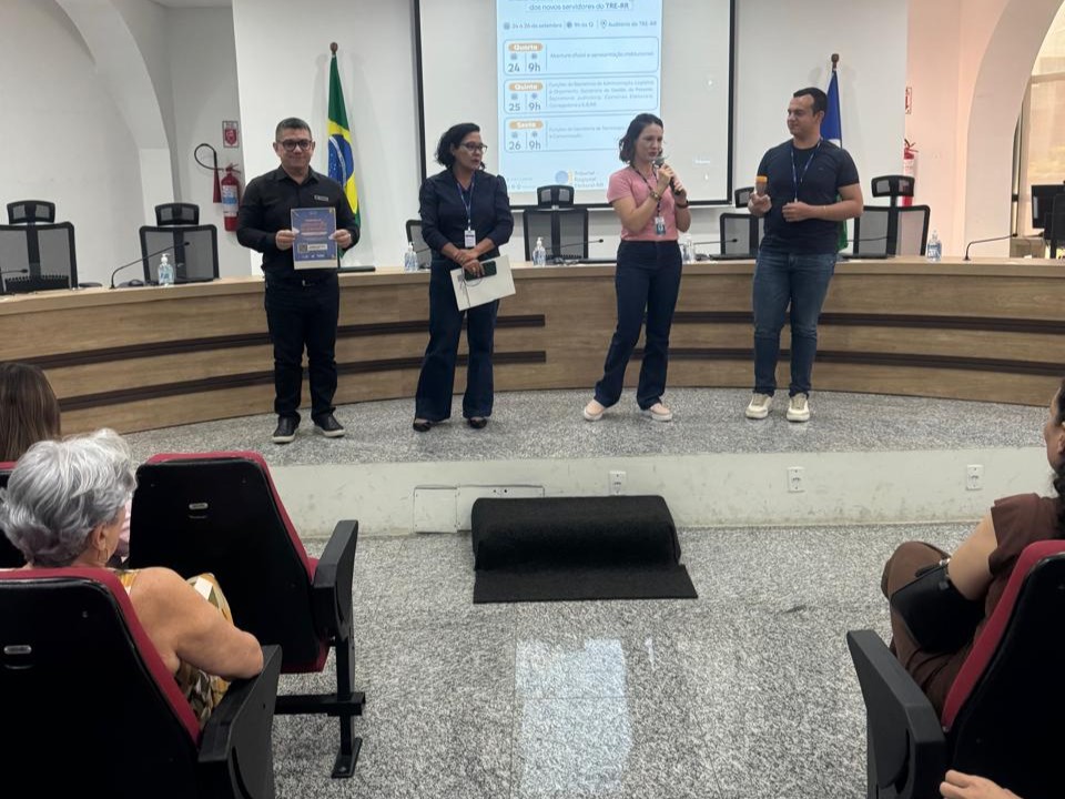 Foto colorida mostra em um auditório. Em pé, no palco, estão quatro servidores, dois homens e duas mulheres. Na frente do palco, pessoas assistem sentadas em cadeiras acolchoadas vermelhas. O piso é de granito claro e o ambiente está bem iluminado com luzes de teto. Foto colorida mostra em um auditório. Em pé, no palco, estão quatro servidores, dois homens e duas mulheres. Na frente do palco, pessoas assistem sentadas em cadeiras acolchoadas vermelhas. O piso é de granito claro e o ambiente está bem iluminado com luzes de teto.