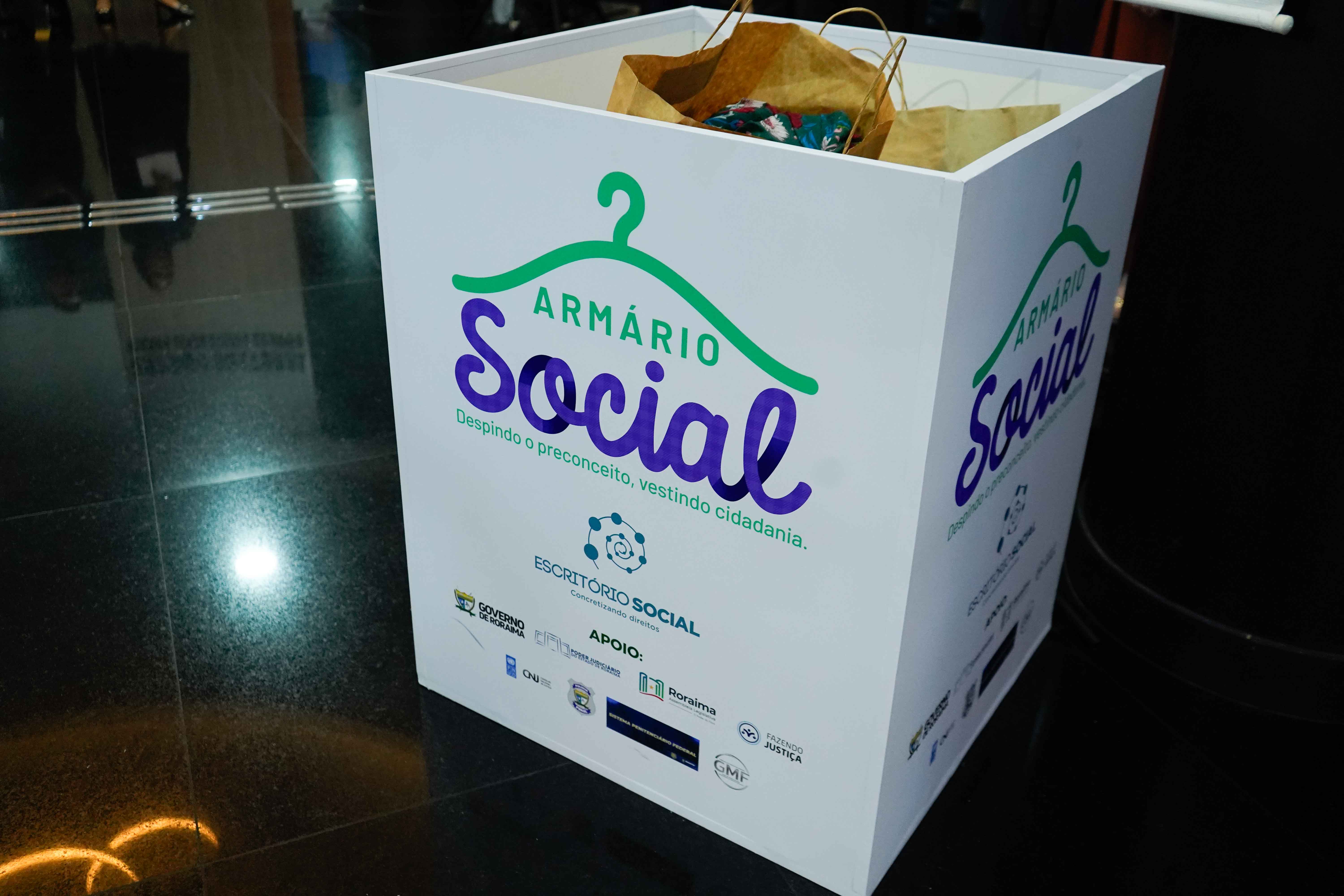 Imagem colorida mostra um primeiro plano de uma grande caixa branca no chão, identificada como "Armário Social" com a frase em português "Despindo o preconceito, vestindo cidadania". A caixa contém alguns sacos de papel e pacotes no topo, sugerindo ser um recipiente de doações. Abaixo do nome do projeto, há logotipos de diversas instituições de apoio em pequenas dimensões. Imagem colorida mostra um primeiro plano de uma grande caixa branca no chão, identificada como "Armário Social" com a frase em português "Despindo o preconceito, vestindo cidadania". A caixa contém alguns sacos de papel e pacotes no topo, sugerindo ser um recipiente de doações. Abaixo do nome do projeto, há logotipos de diversas instituições de apoio em pequenas dimensões.