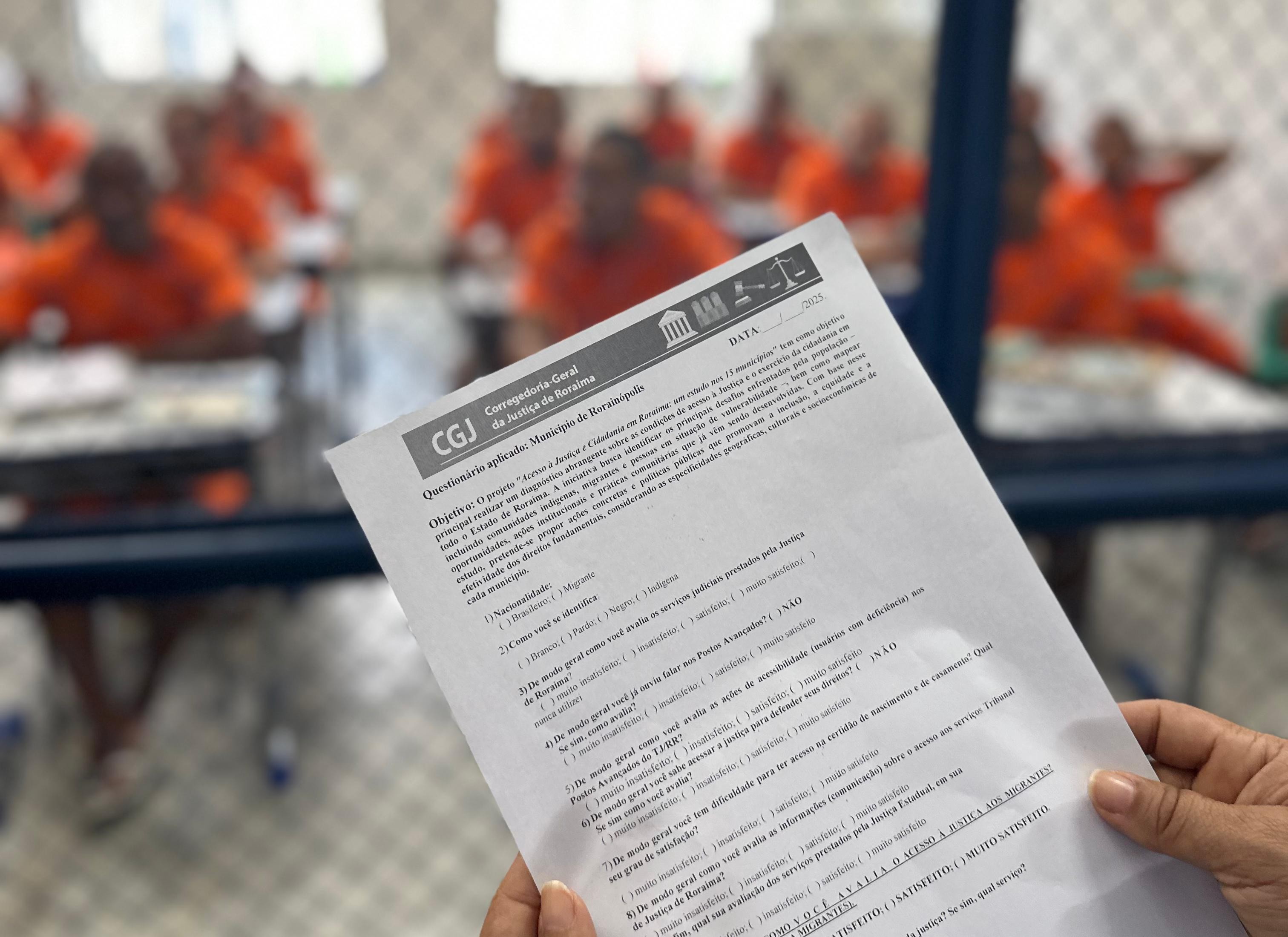 A imagem mostra uma mão segurando um questionário oficial da Corregedoria-Geral da Justiça de Roraima, com o título em destaque e perguntas voltadas à avaliação dos serviços judiciais. Ao fundo, por trás de uma grade metálica, aparecem homens vestidos com uniformes laranja, sentados em carteiras escolares organizadas em fileiras A imagem mostra uma mão segurando um questionário oficial da Corregedoria-Geral da Justiça de Roraima, com o título em destaque e perguntas voltadas à avaliação dos serviços judiciais. Ao fundo, por trás de uma grade metálica, aparecem homens vestidos com uniformes laranja, sentados em carteiras escolares organizadas em fileiras