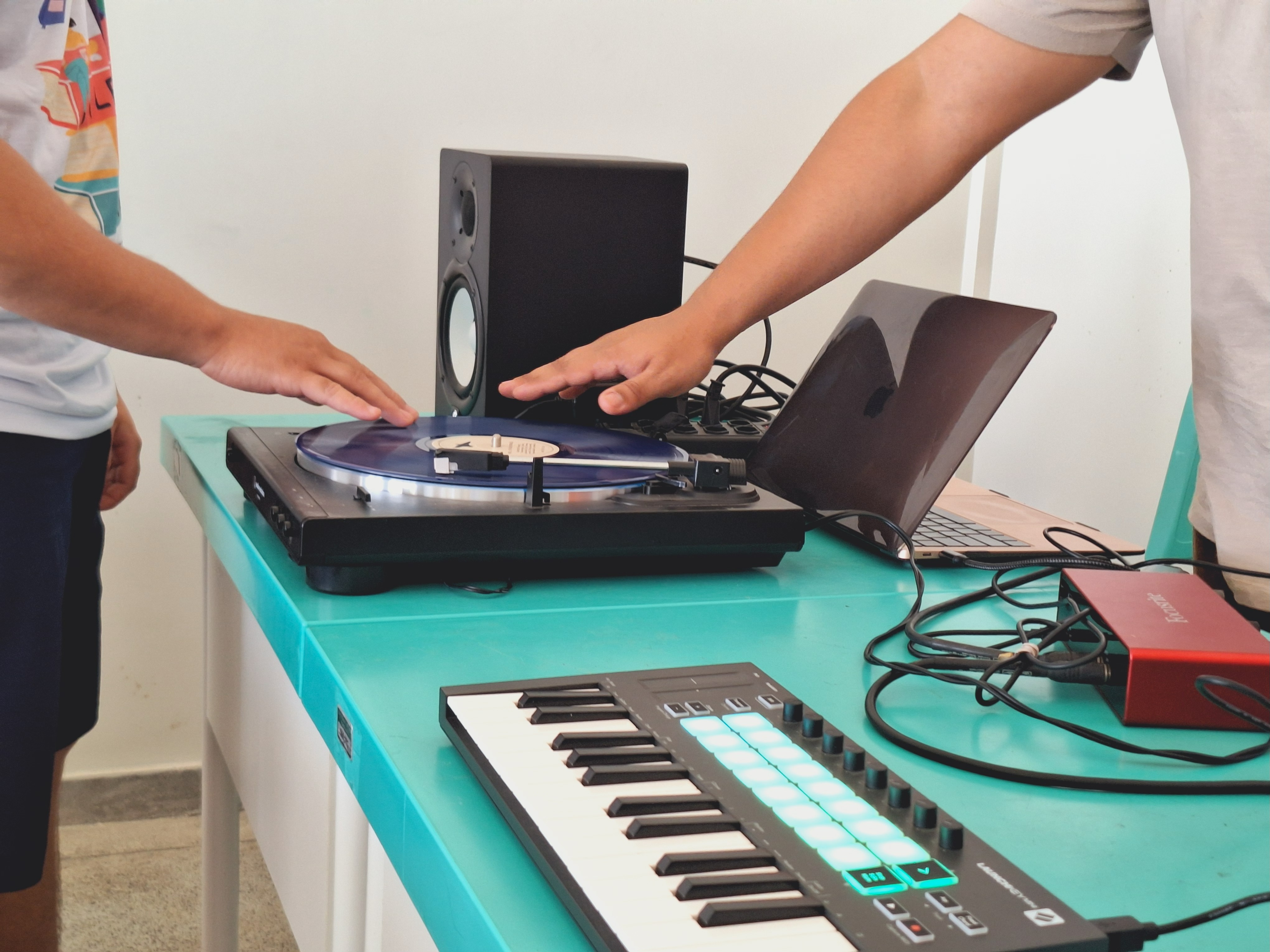 Foto colorida mostra mostra dois jovens interagindo com equipamentos de produção musical sobre uma mesa de cor turquesa. As mãos deles estão posicionadas sobre um toca-discos com um vinil azul. Ao lado, há um teclado com botões iluminados, um laptop aberto, uma caixa de som e uma interface de áudio vermelha com vários cabos conectados. Foto colorida mostra mostra dois jovens interagindo com equipamentos de produção musical sobre uma mesa de cor turquesa. As mãos deles estão posicionadas sobre um toca-discos com um vinil azul. Ao lado, há um teclado com botões iluminados, um laptop aberto, uma caixa de som e uma interface de áudio vermelha com vários cabos conectados.