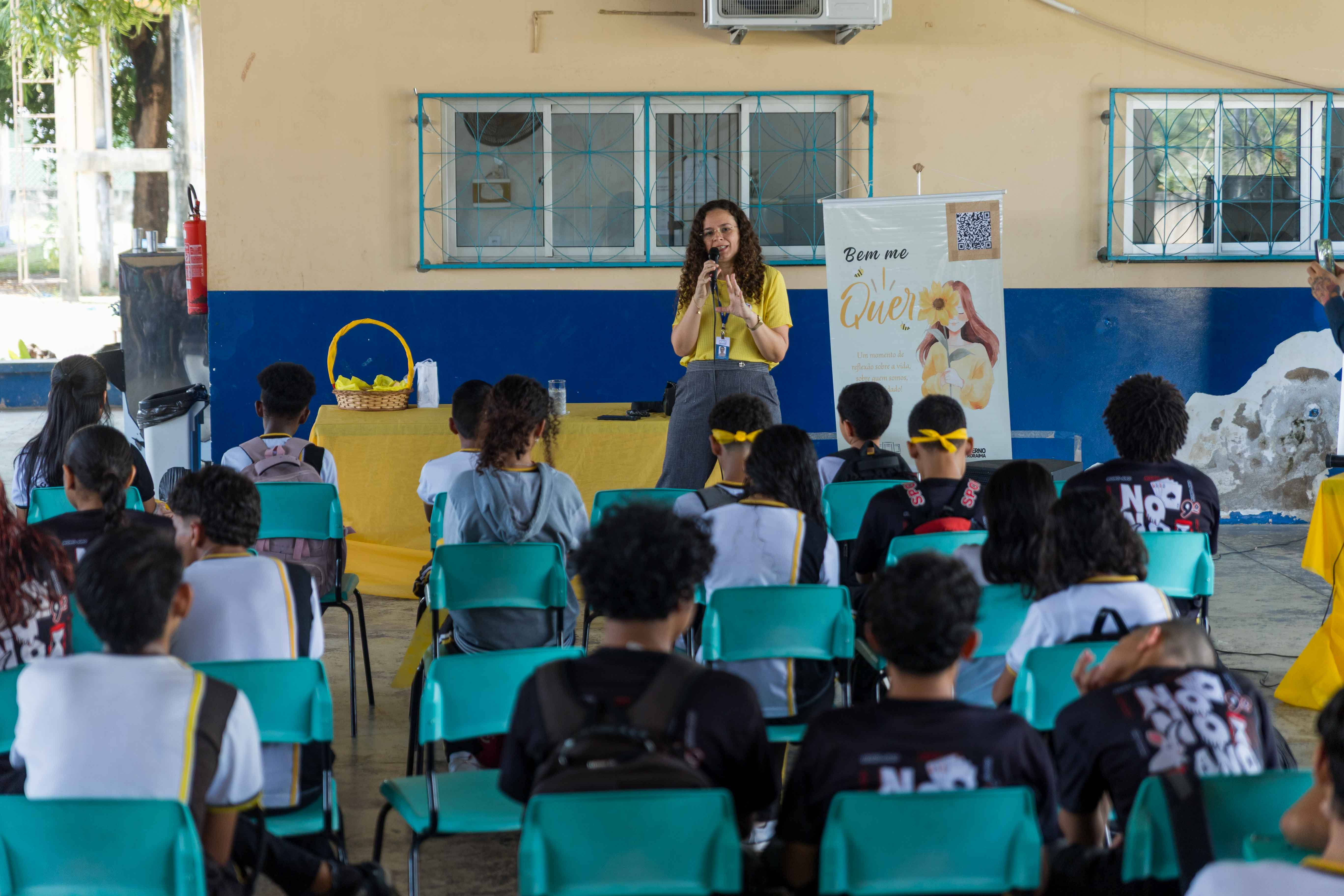 Imagem colorida mostra uma mulher de camisa amarela falando ao microfone para um grupo de jovens sentados em cadeiras azuis. Ao fundo, há um banner amarelo com uma ilustração de girassol e as palavras "Bem me Quero". A cena parece ser uma palestra ou apresentação em um ambiente escolar ou comunitário.