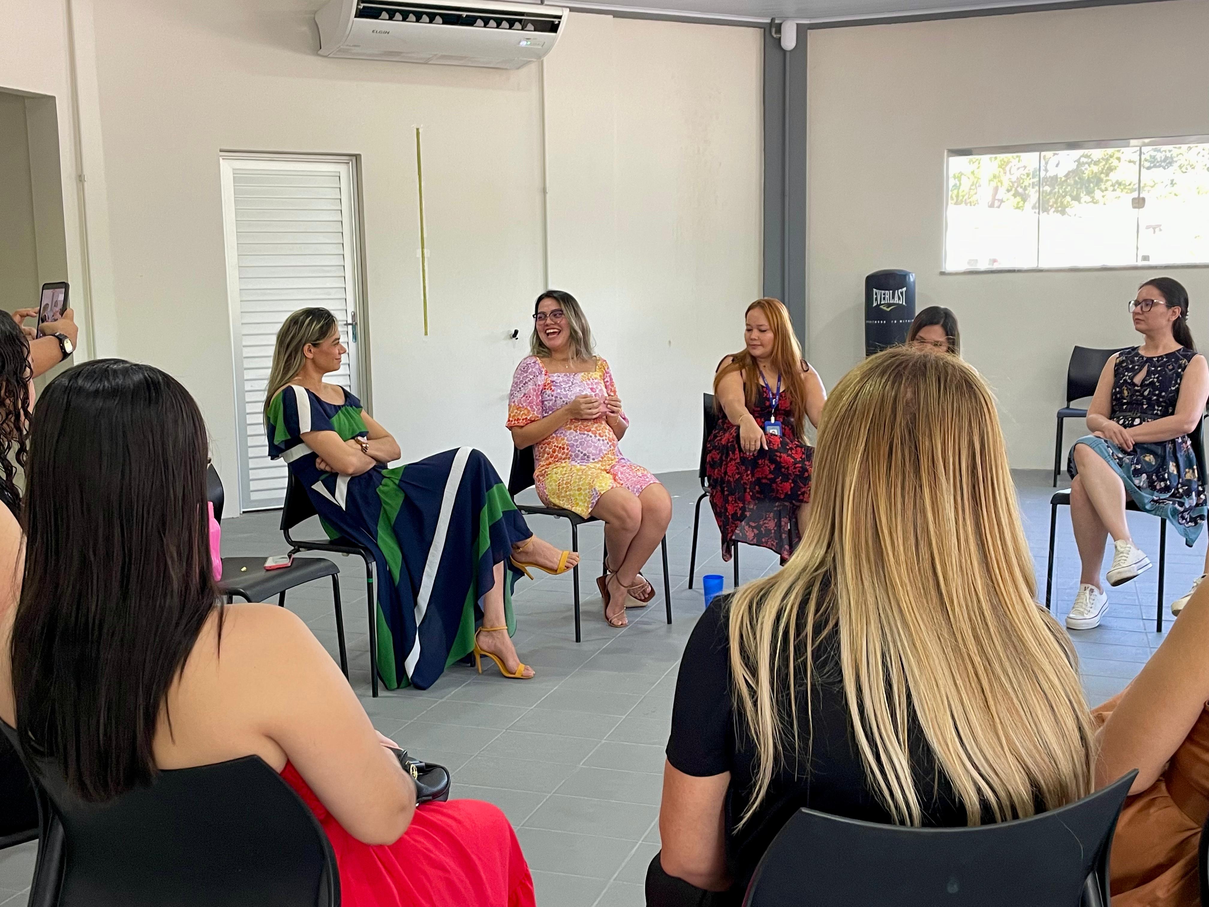A imagem mostra um grupo de mulheres reunidas em círculo dentro de uma sala clara, de paredes brancas e piso cinza. Todas estão sentadas em cadeiras de plástico pretas, posicionadas de forma a favorecer a conversa. Em primeiro plano, vemos mulheres de costas, uma delas com cabelo loiro comprido e outra de vestido vermelho. No centro da imagem, à esquerda, uma delas veste um vestido longo listrado em azul, verde e branco, ao lado, outra mulher sorri enquanto fala, usando um vestido curto estampado em rosa, amarelo e laranja, com sandálias amarelas. À direita, há outras participantes, incluindo uma mulher de vestido preto com flores vermelhas e outra de vestido azul estampado com tênis brancos. No canto esquerdo, alguém segura um celular, registrando o momento. Ao fundo, há uma janela que deixa entrar a luz natural e um aparelho de ar-condicionado fixado na parede. A imagem mostra um grupo de mulheres reunidas em círculo dentro de uma sala clara, de paredes brancas e piso cinza. Todas estão sentadas em cadeiras de plástico pretas, posicionadas de forma a favorecer a conversa. Em primeiro plano, vemos mulheres de costas, uma delas com cabelo loiro comprido e outra de vestido vermelho. No centro da imagem, à esquerda, uma delas veste um vestido longo listrado em azul, verde e branco, ao lado, outra mulher sorri enquanto fala, usando um vestido curto estampado em rosa, amarelo e laranja, com sandálias amarelas. À direita, há outras participantes, incluindo uma mulher de vestido preto com flores vermelhas e outra de vestido azul estampado com tênis brancos. No canto esquerdo, alguém segura um celular, registrando o momento. Ao fundo, há uma janela que deixa entrar a luz natural e um aparelho de ar-condicionado fixado na parede.