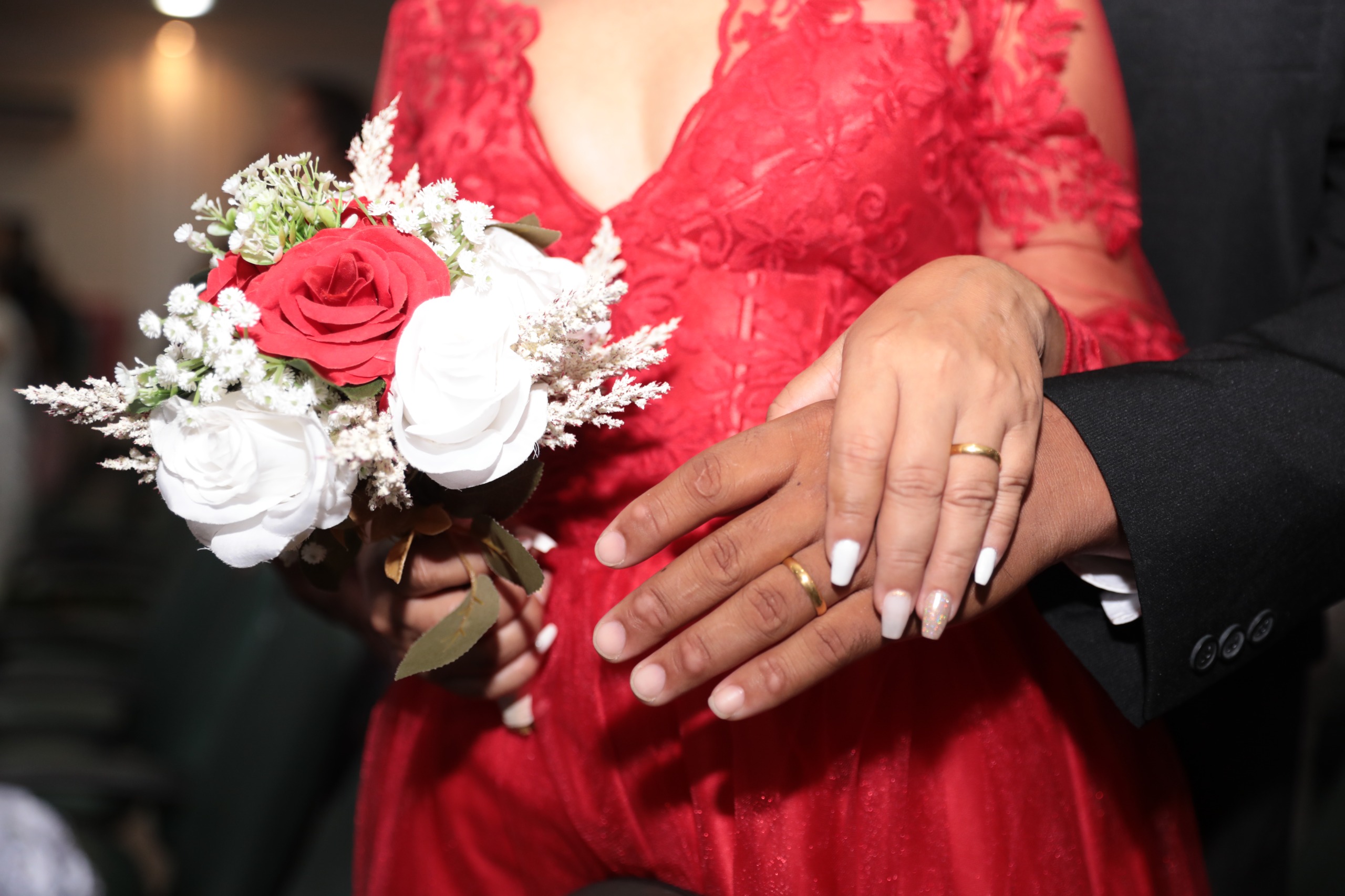 Foto colorida mostra em primeiro plano, um casal de mãos dadas, com as alianças de casamento em destaque. A noiva, com vestido de renda vermelho e unhas pintadas de branco, segura um buquê com rosas vermelhas e brancas. O noivo veste terno preto. Foto colorida mostra em primeiro plano, um casal de mãos dadas, com as alianças de casamento em destaque. A noiva, com vestido de renda vermelho e unhas pintadas de branco, segura um buquê com rosas vermelhas e brancas. O noivo veste terno preto.