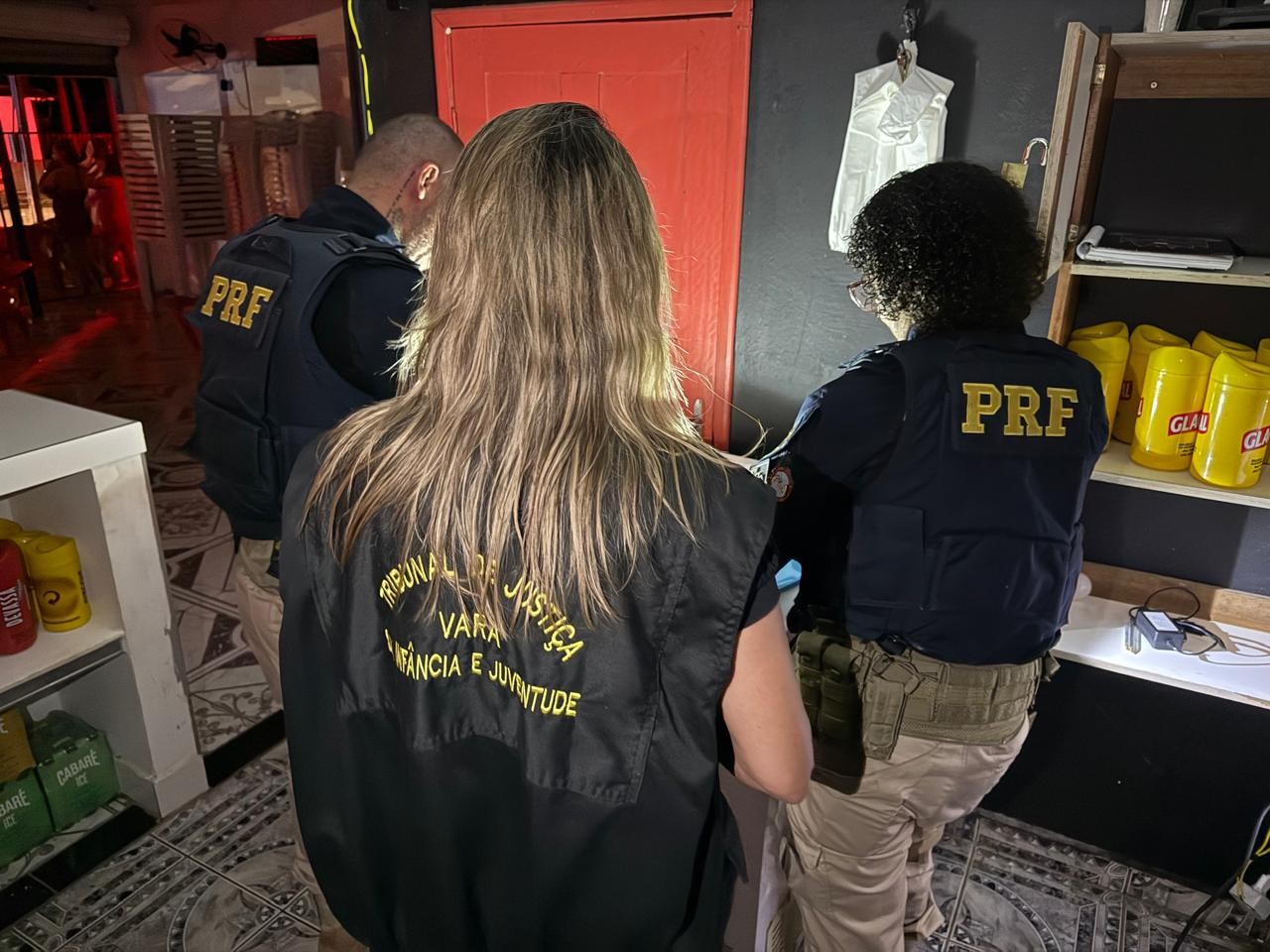A imagem colorida mostra três agentes de fiscalização vistos de costas, em um ambiente interno. Dois deles, nas laterais, vestem coletes da Polícia Rodoviária Federal (PRF). A agente no centro usa um colete preto com a inscrição em amarelo do "Tribunal de Justiça - Vara da Infância e Juventude". A imagem colorida mostra três agentes de fiscalização vistos de costas, em um ambiente interno. Dois deles, nas laterais, vestem coletes da Polícia Rodoviária Federal (PRF). A agente no centro usa um colete preto com a inscrição em amarelo do "Tribunal de Justiça - Vara da Infância e Juventude".