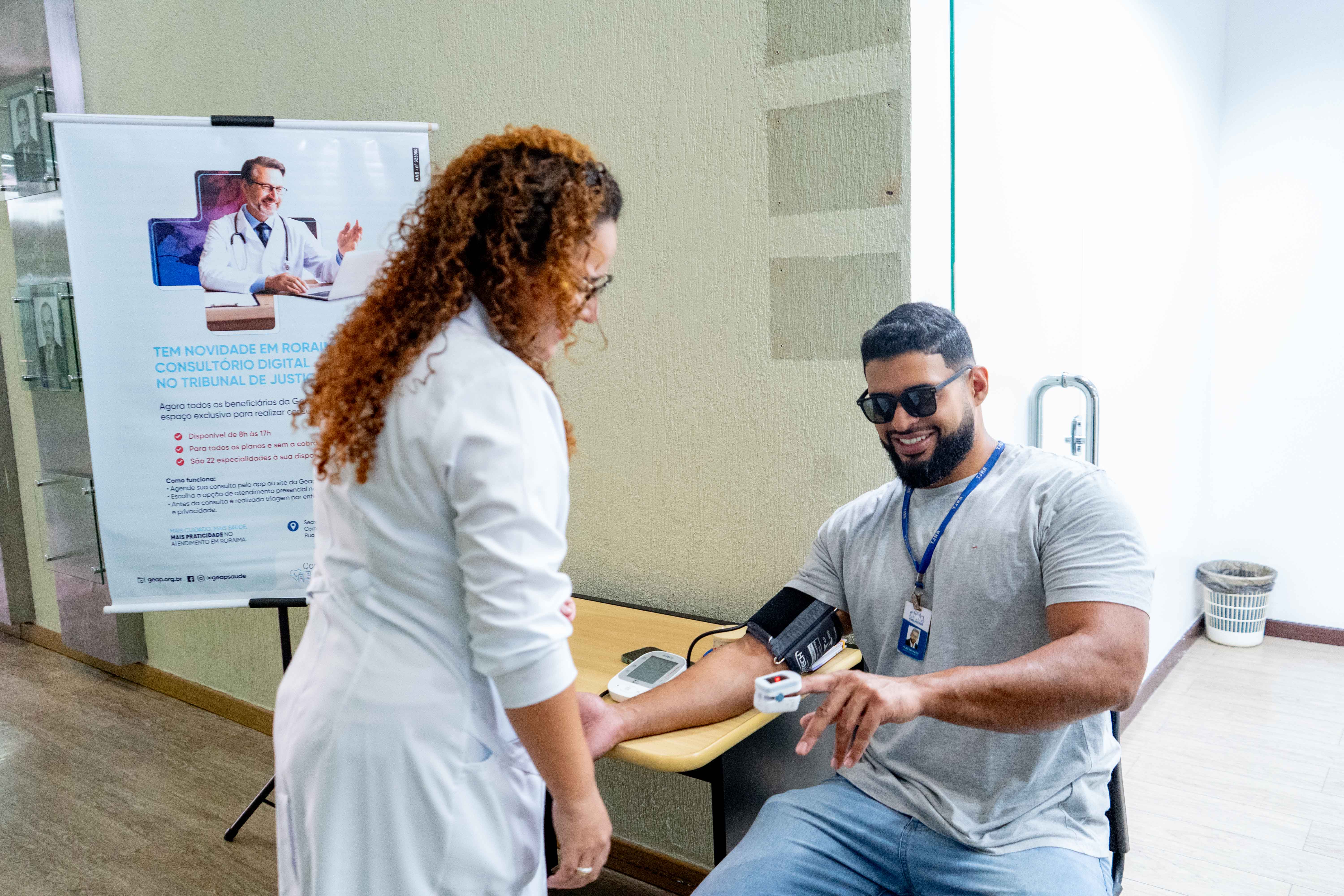 A imagem mostra um ambiente interno onde uma mulher de jaleco branco mede a pressão arterial de um homem com barba, de óculos escuros, que veste uma camisa cinza e está sentado. A mulher está em pé, olhando para o aparelho digital no braço dele, sobre uma mesa de madeira. Ao lado esquerdo, um banner vertical com a imagem de um médico. A imagem mostra um ambiente interno onde uma mulher de jaleco branco mede a pressão arterial de um homem com barba, de óculos escuros, que veste uma camisa cinza e está sentado. A mulher está em pé, olhando para o aparelho digital no braço dele, sobre uma mesa de madeira. Ao lado esquerdo, um banner vertical com a imagem de um médico.