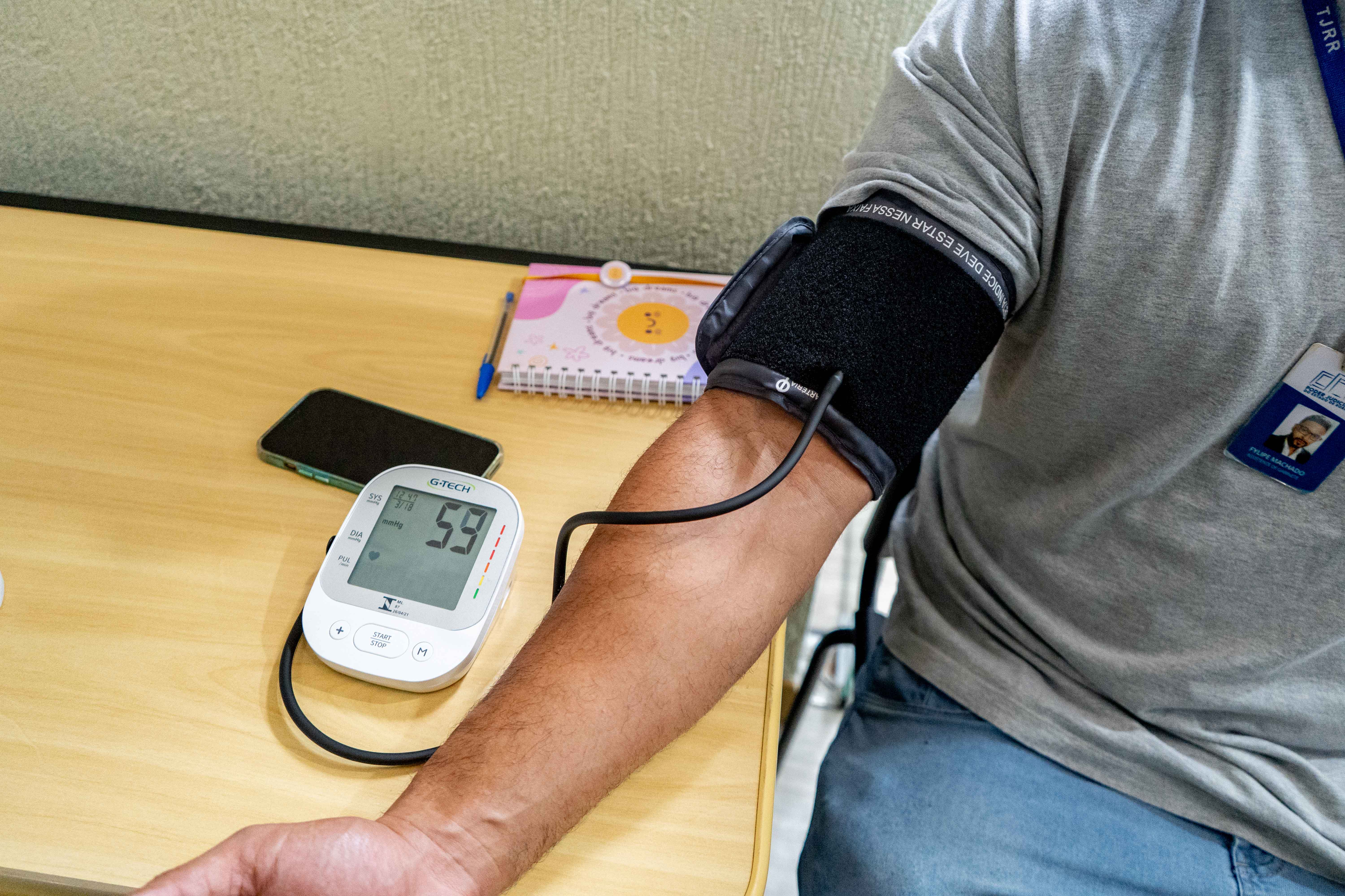A imagem mostra um braço de um homem, no qual um aparelho digital está medindo sua pressão arterial. O monitor do aparelho está sobre uma mesa de madeira clara e exibe a leitura do pulso e os valores de pressão arterial. O homem veste uma camisa cinza e calça jeans azul. Sobre a mesa, há também um celular preto e um caderno de anotações. A imagem mostra um braço de um homem, no qual um aparelho digital está medindo sua pressão arterial. O monitor do aparelho está sobre uma mesa de madeira clara e exibe a leitura do pulso e os valores de pressão arterial. O homem veste uma camisa cinza e calça jeans azul. Sobre a mesa, há também um celular preto e um caderno de anotações.