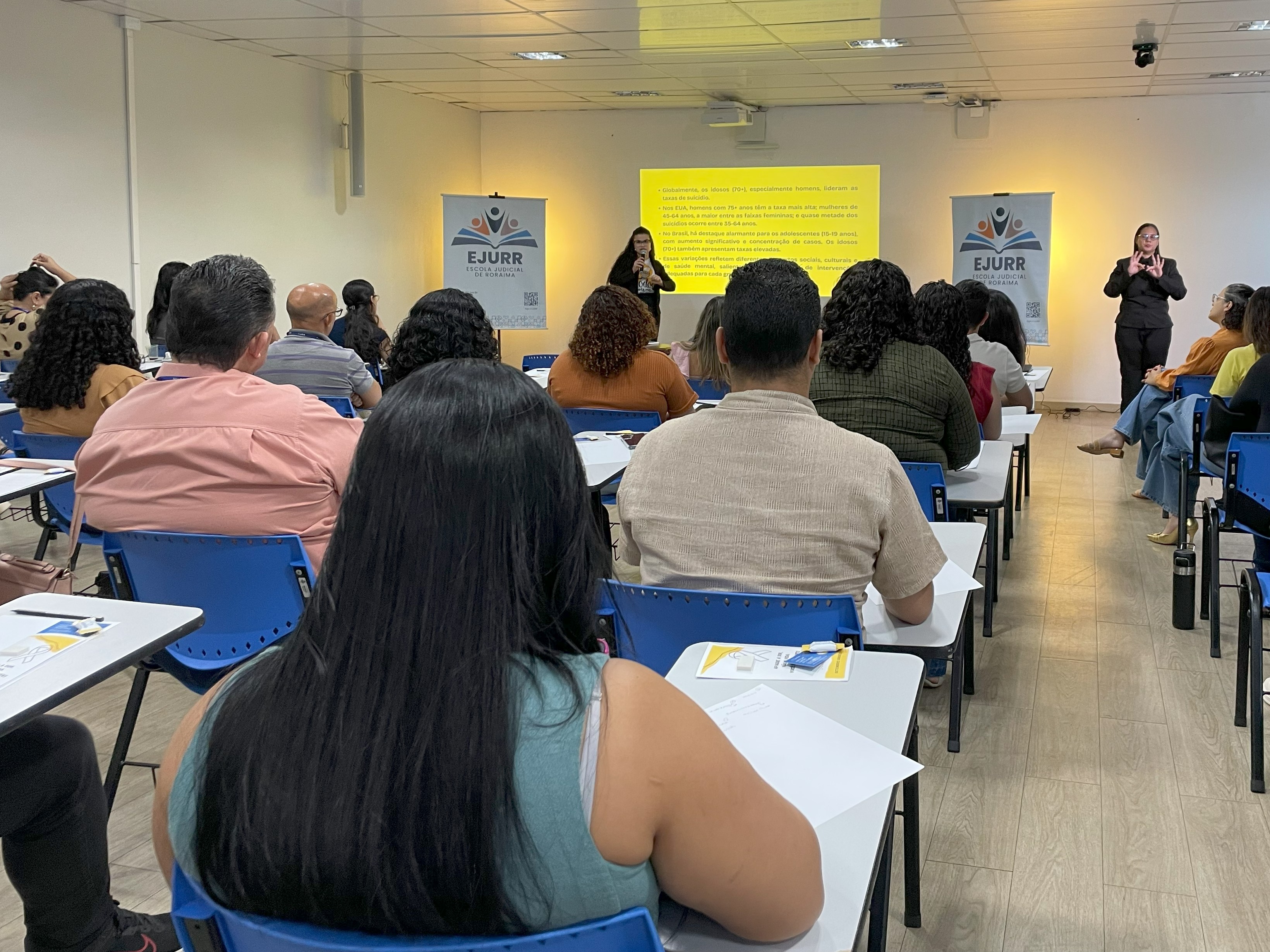 A imagem colorida mostra uma sala, com participantes sentados em cadeiras escolares azuis, voltados para o fundo da sala, onde ocorre uma palestra. No centro do ambiente, uma mulher fala ao microfone, enquanto há uma do outro lado fazendo libras. A imagem colorida mostra uma sala, com participantes sentados em cadeiras escolares azuis, voltados para o fundo da sala, onde ocorre uma palestra. No centro do ambiente, uma mulher fala ao microfone, enquanto há uma do outro lado fazendo libras.