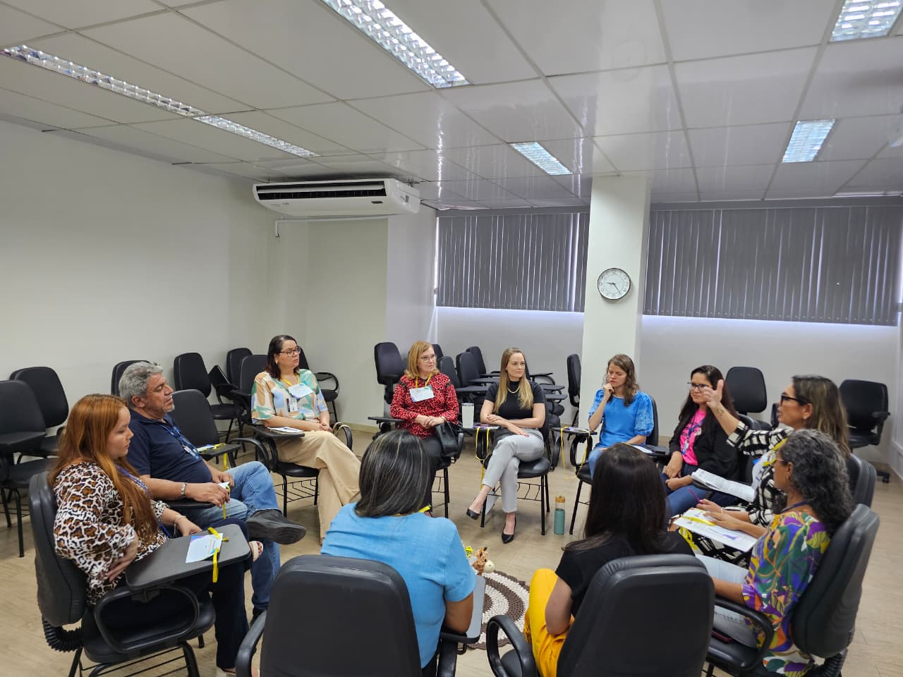 A imagem colorida de um grupo de dez adultos (oito mulheres e dois homens) está sentado em cadeiras de escritório pretas com pranchetas acopladas, formando um círculo informal para facilitar a discussão. A imagem colorida de um grupo de dez adultos (oito mulheres e dois homens) está sentado em cadeiras de escritório pretas com pranchetas acopladas, formando um círculo informal para facilitar a discussão.