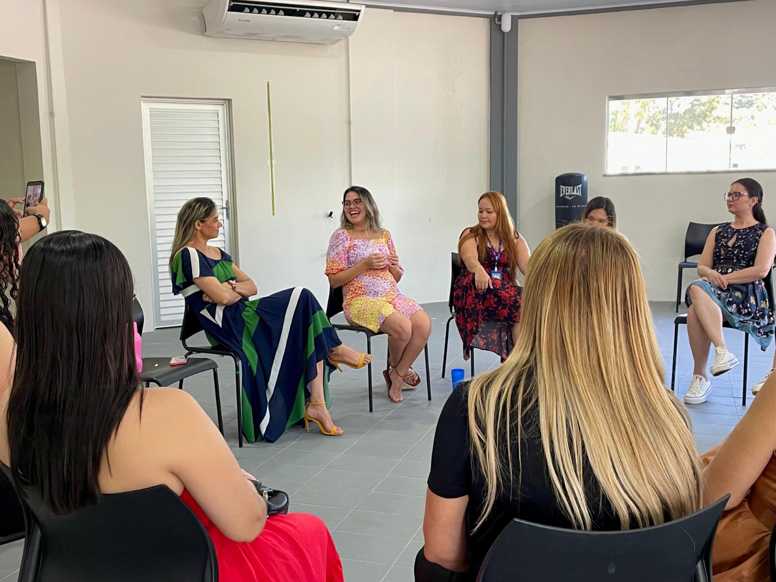 A imagem mostra um grupo de mulheres reunidas em círculo dentro de uma sala clara, de paredes brancas e piso cinza. Todas estão sentadas em cadeiras de plástico pretas, posicionadas de forma a favorecer a conversa. A imagem mostra um grupo de mulheres reunidas em círculo dentro de uma sala clara, de paredes brancas e piso cinza. Todas estão sentadas em cadeiras de plástico pretas, posicionadas de forma a favorecer a conversa.