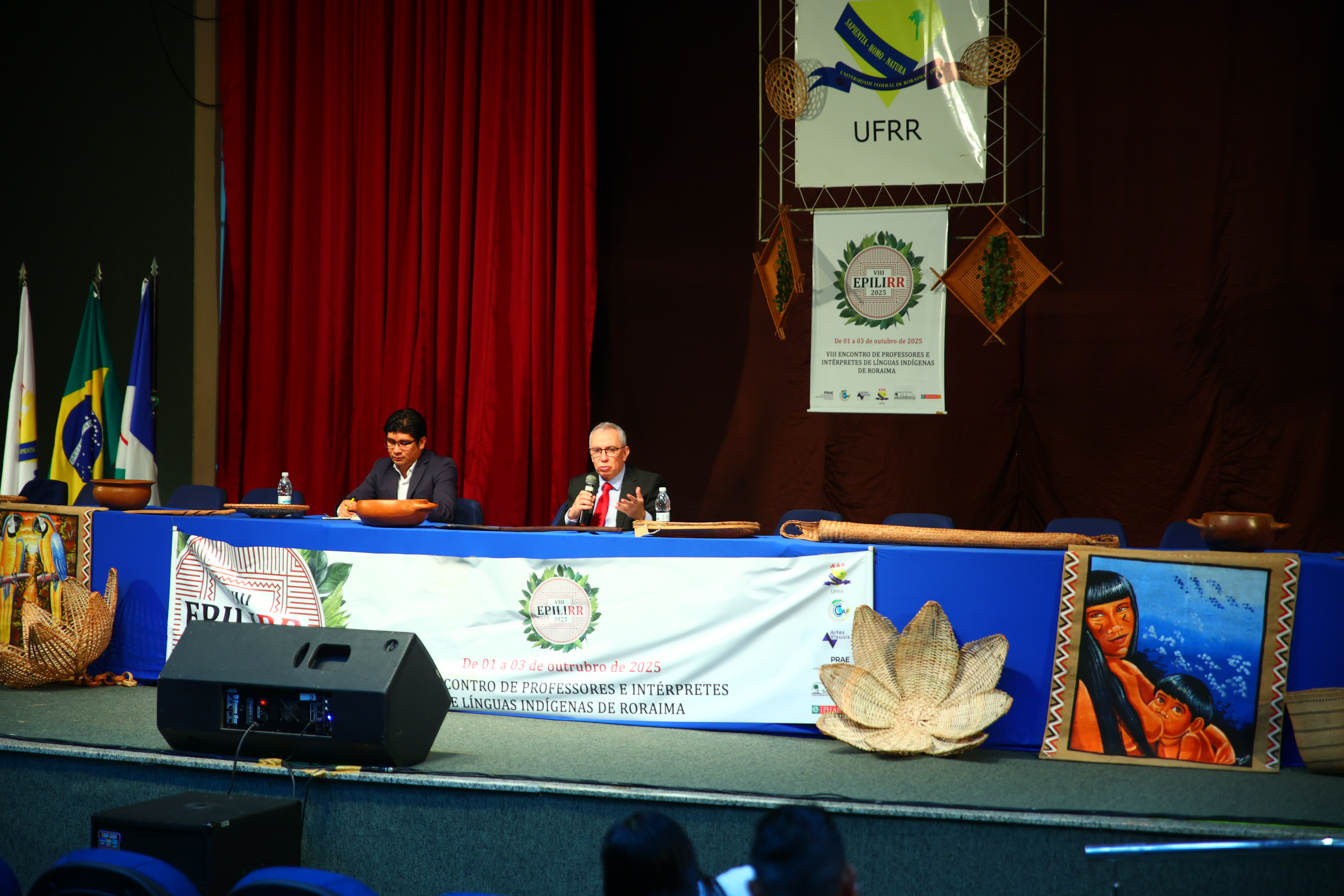 Imagem colorida mostra uma palestra em auditório. Os dois homens sentados na mesa de palco azul, um falando ao microfone, que é o desembargador Almiro Padilha, Vice-Presidente do Tribunal de Justiça de Roraima são visíveis. Há banners com logotipos, junto com uma cortina vermelha e objetos de artesanato indígena no palco.