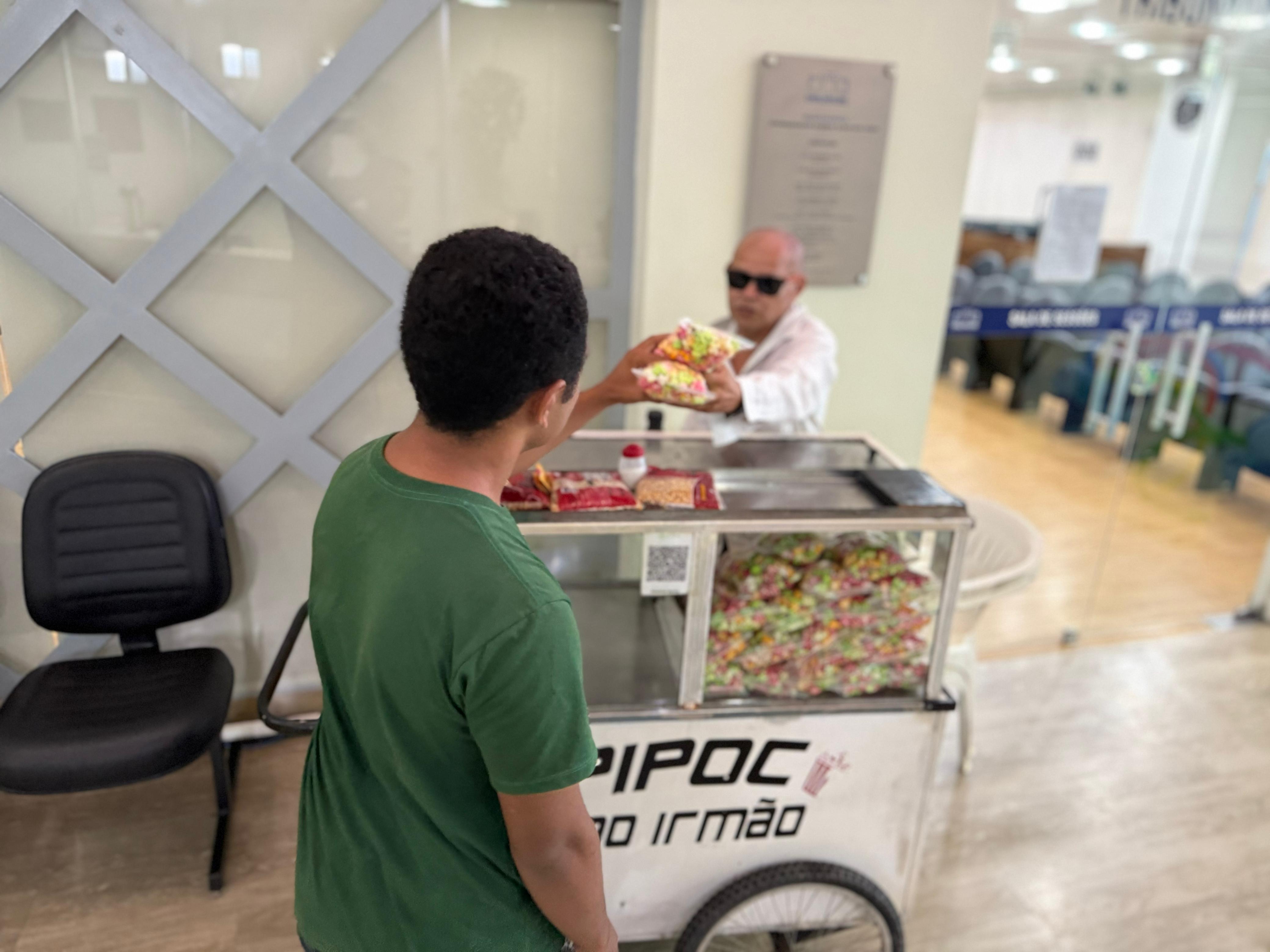 Imagem colorida mostra um carrinho de pipoca sendo operado por um homem de óculos escuros, que está entregando um saco de pipoca colorida para uma pessoa de costas, vestindo uma camiseta verde. O carrinho, de cor branca com rodas e o nome "Pipoca do Irmão" visível na frente, está repleto de sacos de pipoca colorida. A cena se passa em um ambiente interno e iluminado, com uma cadeira escura à esquerda e uma parede com painéis de vidro e metal ao fundo. Imagem colorida mostra um carrinho de pipoca sendo operado por um homem de óculos escuros, que está entregando um saco de pipoca colorida para uma pessoa de costas, vestindo uma camiseta verde. O carrinho, de cor branca com rodas e o nome "Pipoca do Irmão" visível na frente, está repleto de sacos de pipoca colorida. A cena se passa em um ambiente interno e iluminado, com uma cadeira escura à esquerda e uma parede com painéis de vidro e metal ao fundo.
