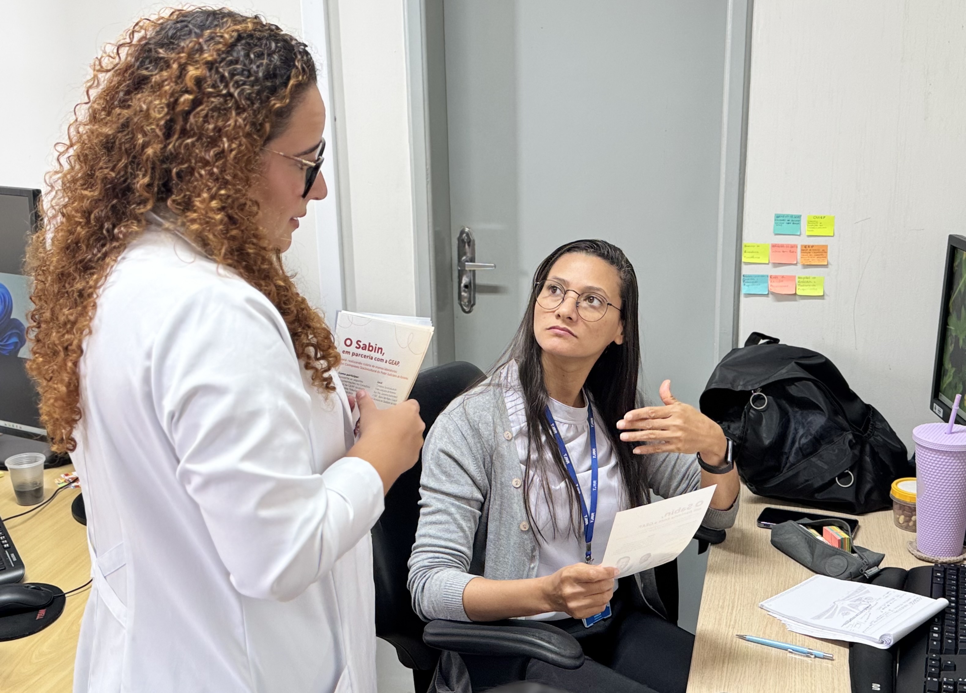 Foto colorida mostra à esquerda, uma profissional de jaleco branco e cabelos cacheados segurando panfletos informativos do Sabin e da GEAP está de pé, dialogando com a outra mulher, que está sentada em frente a uma mesa. A mulher sentada usa óculos, crachá azul e veste camiseta branca com cardigã cinza claro. Ela segura um dos panfletos, escutando com atenção, enquanto gesticula com a mão livre. Foto colorida mostra à esquerda, uma profissional de jaleco branco e cabelos cacheados segurando panfletos informativos do Sabin e da GEAP está de pé, dialogando com a outra mulher, que está sentada em frente a uma mesa. A mulher sentada usa óculos, crachá azul e veste camiseta branca com cardigã cinza claro. Ela segura um dos panfletos, escutando com atenção, enquanto gesticula com a mão livre.