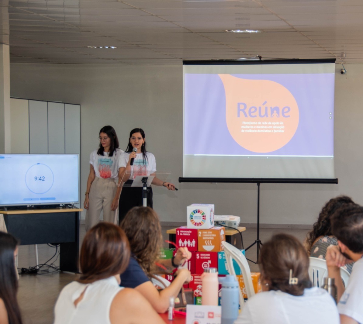 Imagem colorida de duas mulheres realizando uma apresentação. O local é um espaço fechado, mas bem amplo. Há também um slide de contéudo da apresentação escrito "reúne". Imagem colorida de duas mulheres realizando uma apresentação. O local é um espaço fechado, mas bem amplo. Há também um slide de contéudo da apresentação escrito "reúne".