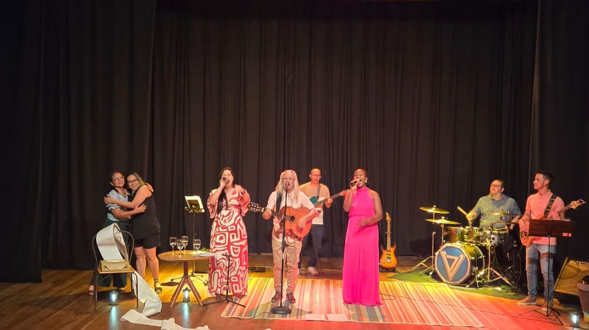 Imagem colorida mostra um palco de teatro com cortinas pretas ao fundo, iluminado por luzes amarelas e coloridas. No centro, uma banda se apresenta. Três vocalistas estão à frente, em microfones: uma mulher de vestido longo estampado em branco e vermelho, um homem de cabelos brancos tocando violão e uma mulher de vestido longo rosa choque. Atrás, estão um guitarrista, um baterista e um baixista. No canto esquerdo, duas mulheres se abraçam perto de uma mesa. O chão de madeira está coberto por um tapete listrado nas cores verde, amarelo e vermelho.