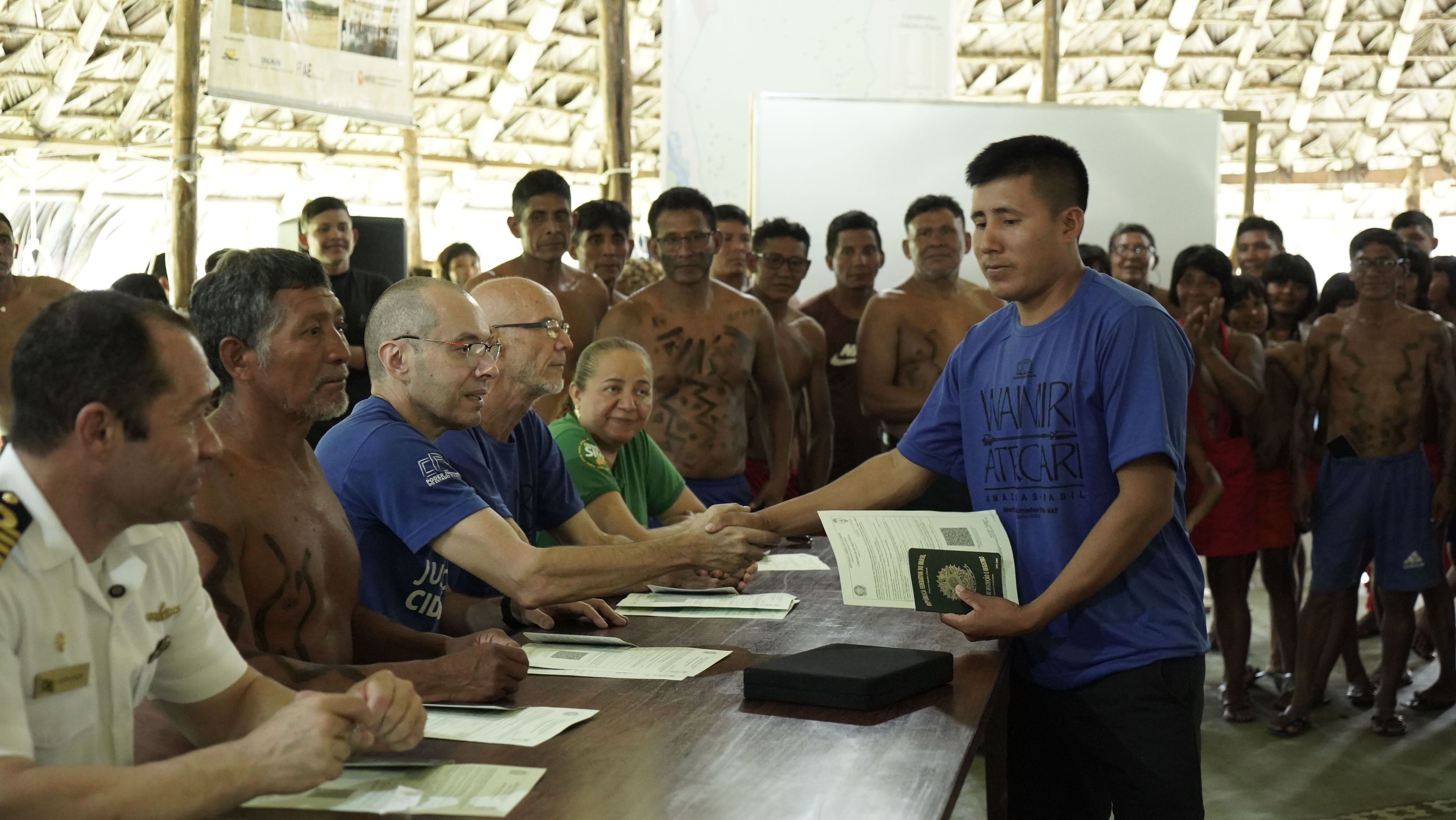 Em uma cerimônia realizada em uma comunitária indígena, um homem recebe seu certificado das mãos de autoridades e representantes do projeto, enquanto outros integrantes da comunidade observam e aplaudem ao fundo.