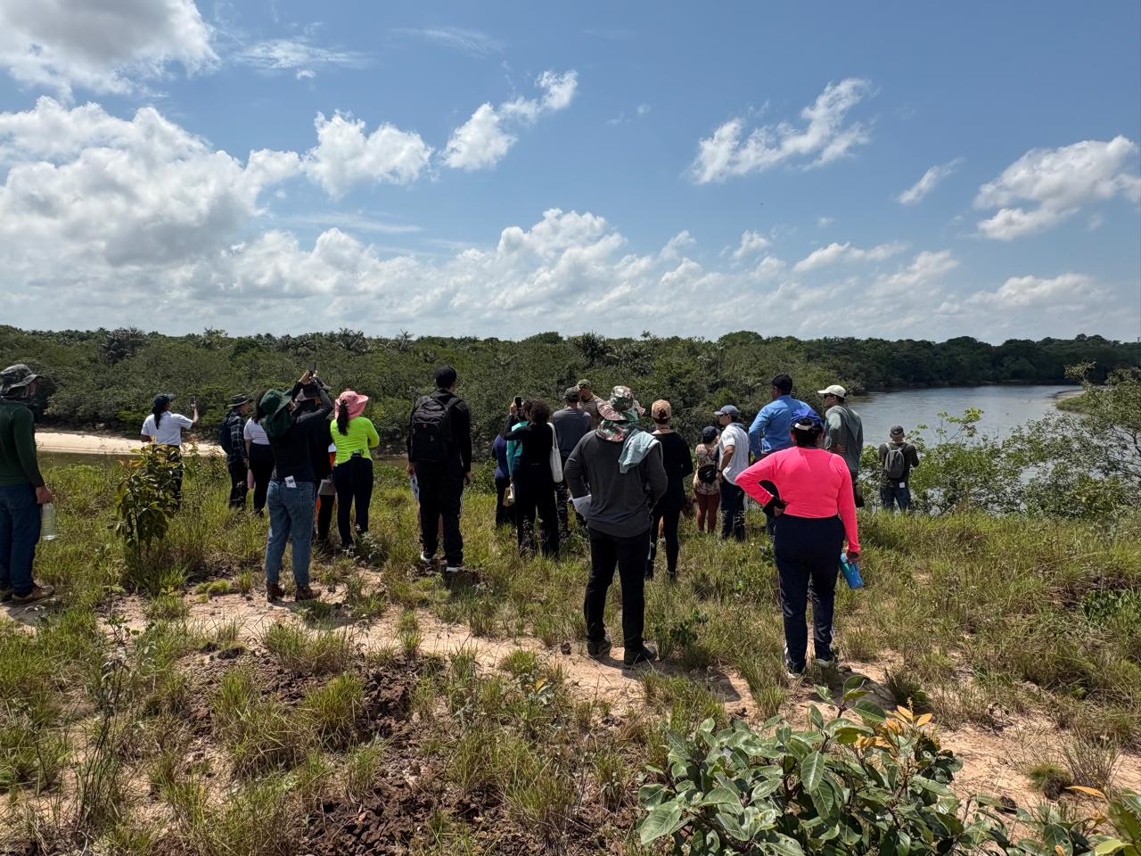 Uma foto tirada ao ar livre em frente ao grupo, há uma paisagem de mata fechada e, ao fundo, é possível ver um rio ou lago cercado por árvores. O céu está azul com nuvens brancas. A cena sugere uma atividade de ecoturismo, estudo da natureza ou passeio em área ribeirinha.