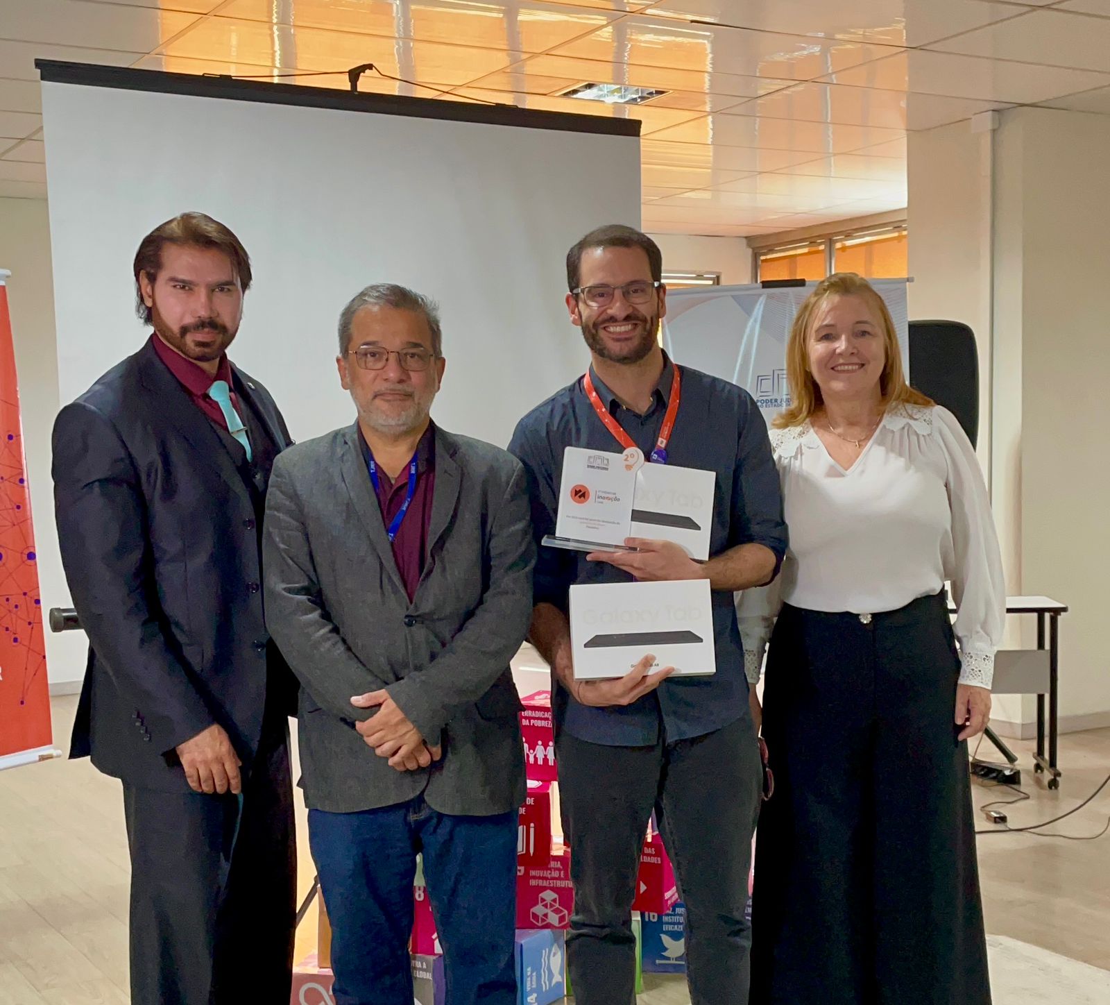 Imagem colorida mostra quatro pessoas posando para uma foto em ambiente interno, com fundo de telão branco e banners institucionais. À direita, está a Desembargadora Elaine Bianchi, usando blusa branca e sorrindo. Ao seu lado, o servidor Tiago Lobo segura duas caixas de Galaxy Tab e um troféu de premiação. À esquerda de Tiago, está o secretário-geral do TJRR, Hermenegildo D’Ávila, vestindo blazer cinza e óculos.