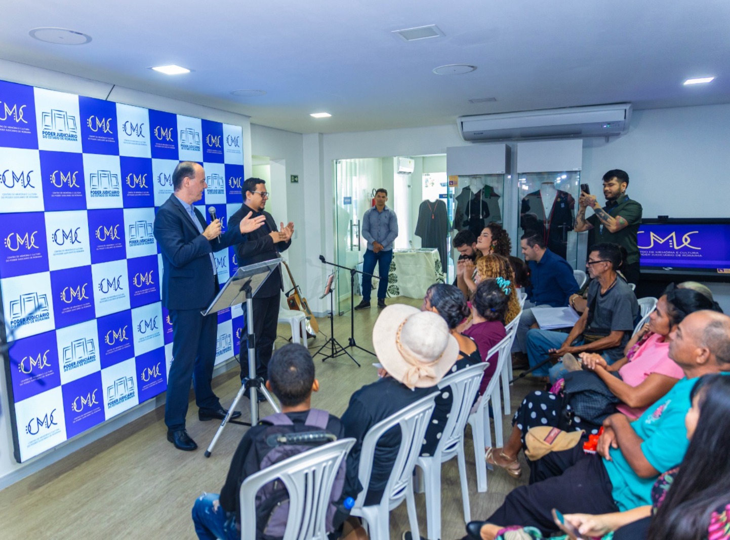 Uma apresentação ou discurso está ocorrendo em um espaço interno. Dois homens estão em frente a um backdrop azul e branco com um logo que inclui as letras "CMC" (e possivelmente "Roraima" ou outro texto menor), falando para uma plateia sentada em cadeiras brancas. Há equipamentos de áudio (microfones) e vídeo (uma TV lateral exibindo o mesmo logo).