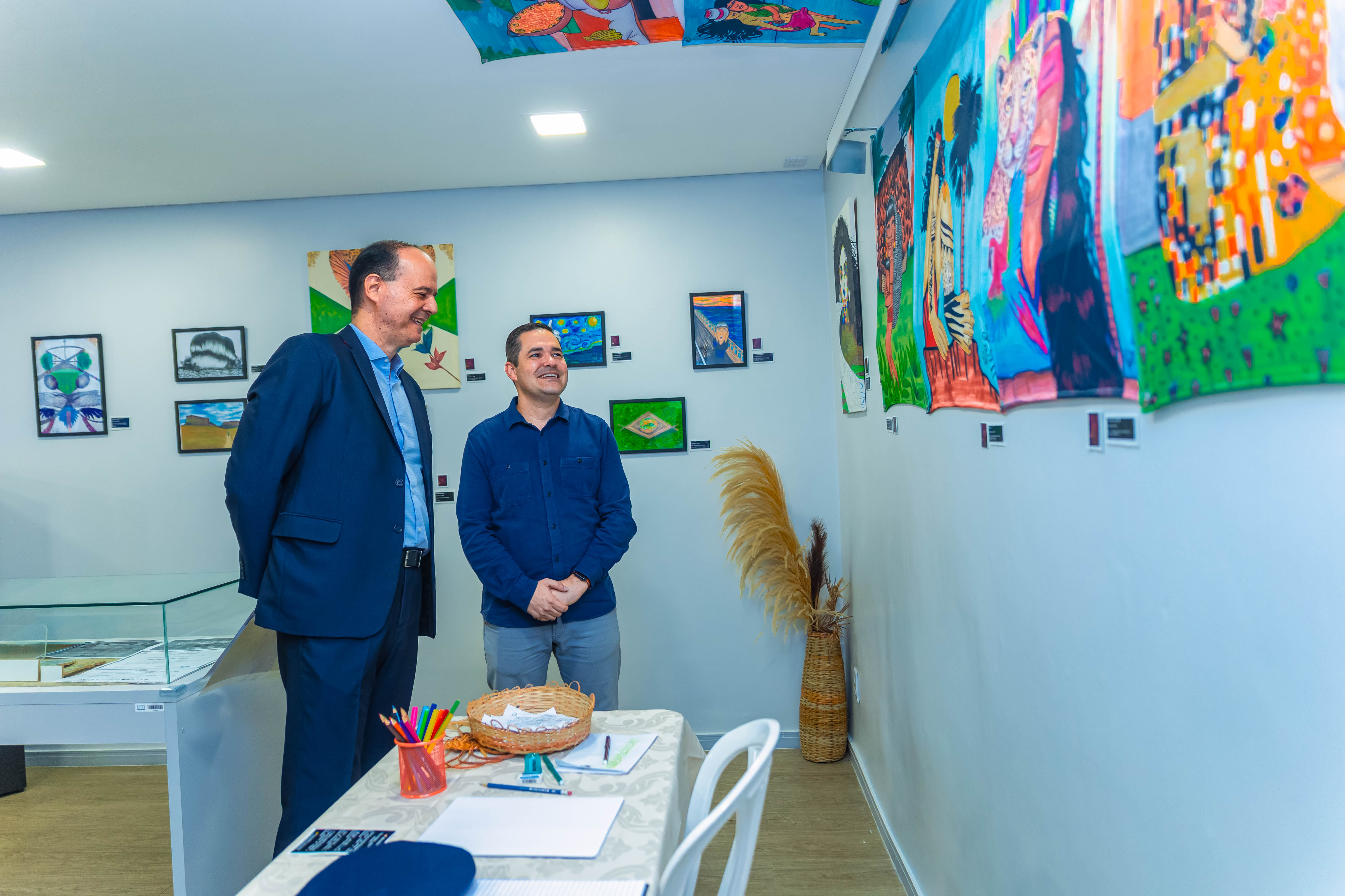 Imagem colorida do  desembargador Cristóvão Suter e do Subcoordenador do CMC, Felipe Queiroz, sorriem enquanto observam pinturas coloridas penduradas em uma parede branca. Há também quadros menores emoldurados e uma mesa com materiais de desenho (lápis de cor) e um cesto.