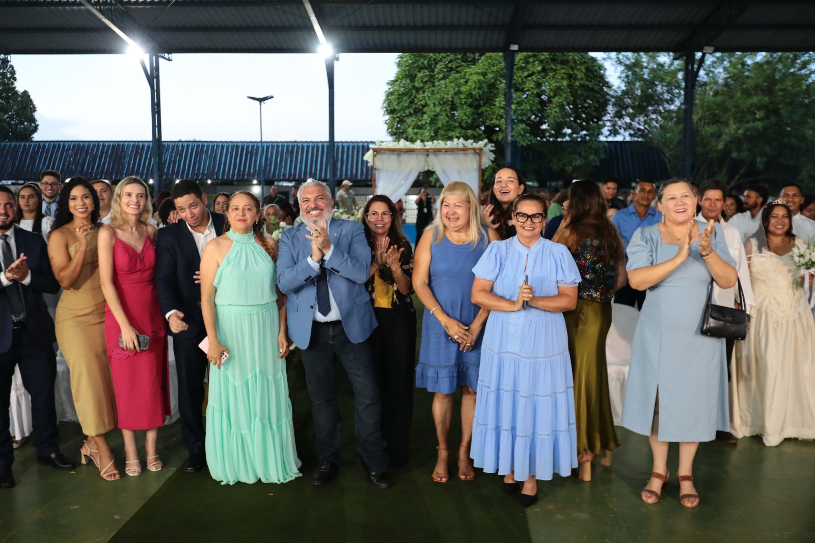 Imagem colorida mostra um grupo de pessoas em um evento social. No centro, um homem de paletó azul escuro e camisa social azul clara sorri e bate palmas. Ele está ladeado por mulheres e outros homens em trajes formais. A mulher mais à frente no centro veste um vestido azul claro longo e rodado, com óculos. À sua esquerda, uma mulher em um vestido azul-acinzentado longo bate palmas, e à direita do homem de paletó, uma mulher em vestido azul vibrante bate palmas. Pessoas em trajes formais, incluindo uma mulher de vestido vermelho vibrante, estão à esquerda. O grupo está sob uma cobertura, com o fundo apresentando uma estrutura decorada com folhagens e flores. Imagem colorida mostra um grupo de pessoas em um evento social. No centro, um homem de paletó azul escuro e camisa social azul clara sorri e bate palmas. Ele está ladeado por mulheres e outros homens em trajes formais. A mulher mais à frente no centro veste um vestido azul claro longo e rodado, com óculos. À sua esquerda, uma mulher em um vestido azul-acinzentado longo bate palmas, e à direita do homem de paletó, uma mulher em vestido azul vibrante bate palmas. Pessoas em trajes formais, incluindo uma mulher de vestido vermelho vibrante, estão à esquerda. O grupo está sob uma cobertura, com o fundo apresentando uma estrutura decorada com folhagens e flores.