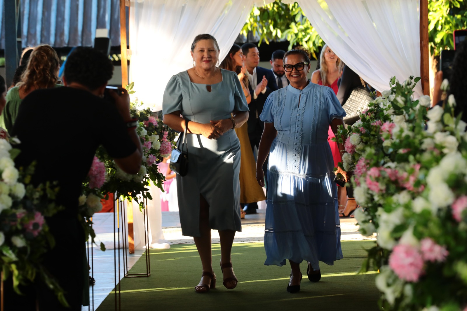 Imagem colorida mostra duas mulheres caminhando em direção à câmera em um evento formal, atravessando um corredor decorado com cortinas brancas e arranjos florais com tons de rosa, branco e verde. A mulher à esquerda veste um vestido azul-acinzentado de comprimento médio com fenda frontal e manga bufante curta, e carrega uma pequena bolsa preta. A mulher à direita veste um vestido longo azul claro com detalhes em renda, e usa óculos. Ambas sorriem. Um fotógrafo está no canto inferior esquerdo, de costas para a imagem. Outras pessoas estão no fundo, atrás das duas mulheres.