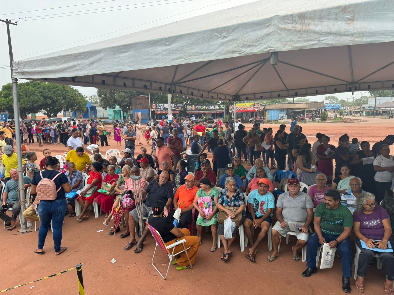  Imagem colorida mostra uma grande concentração de pessoas reunidas sob uma tenda branca montada em um espaço aberto com chão de terra vermelha. Pessoas idosas formam a maioria no primeiro plano e no centro, sentadas em cadeiras plásticas brancas e coloridas. Há uma variedade de cores de roupas. Muitas pessoas estão em pé atrás e nas laterais da área de assentos. 