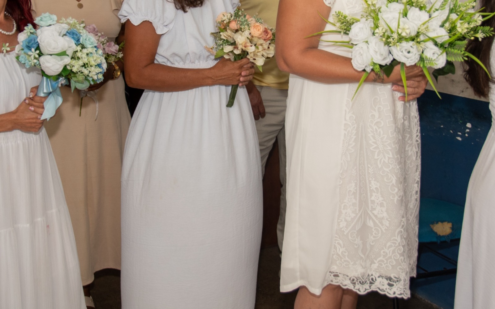 imagem em plano médio de um grupo de pessoas em pé, focada da cintura para baixo, sugerindo uma cerimônia de casamento. Pelo menos três mulheres estão vestindo roupas brancas, semelhantes a vestidos de noiva , e seguram buquês de flores em tons de branco, azul claro e pêssego. Uma das mulheres, no centro-direita, veste um vestido curto com renda na barra e segura um buquê grande de rosas brancas e folhagens. Ao fundo, outras pessoas e um assento azul escuro são visíveis. imagem em plano médio de um grupo de pessoas em pé, focada da cintura para baixo, sugerindo uma cerimônia de casamento. Pelo menos três mulheres estão vestindo roupas brancas, semelhantes a vestidos de noiva , e seguram buquês de flores em tons de branco, azul claro e pêssego. Uma das mulheres, no centro-direita, veste um vestido curto com renda na barra e segura um buquê grande de rosas brancas e folhagens. Ao fundo, outras pessoas e um assento azul escuro são visíveis.