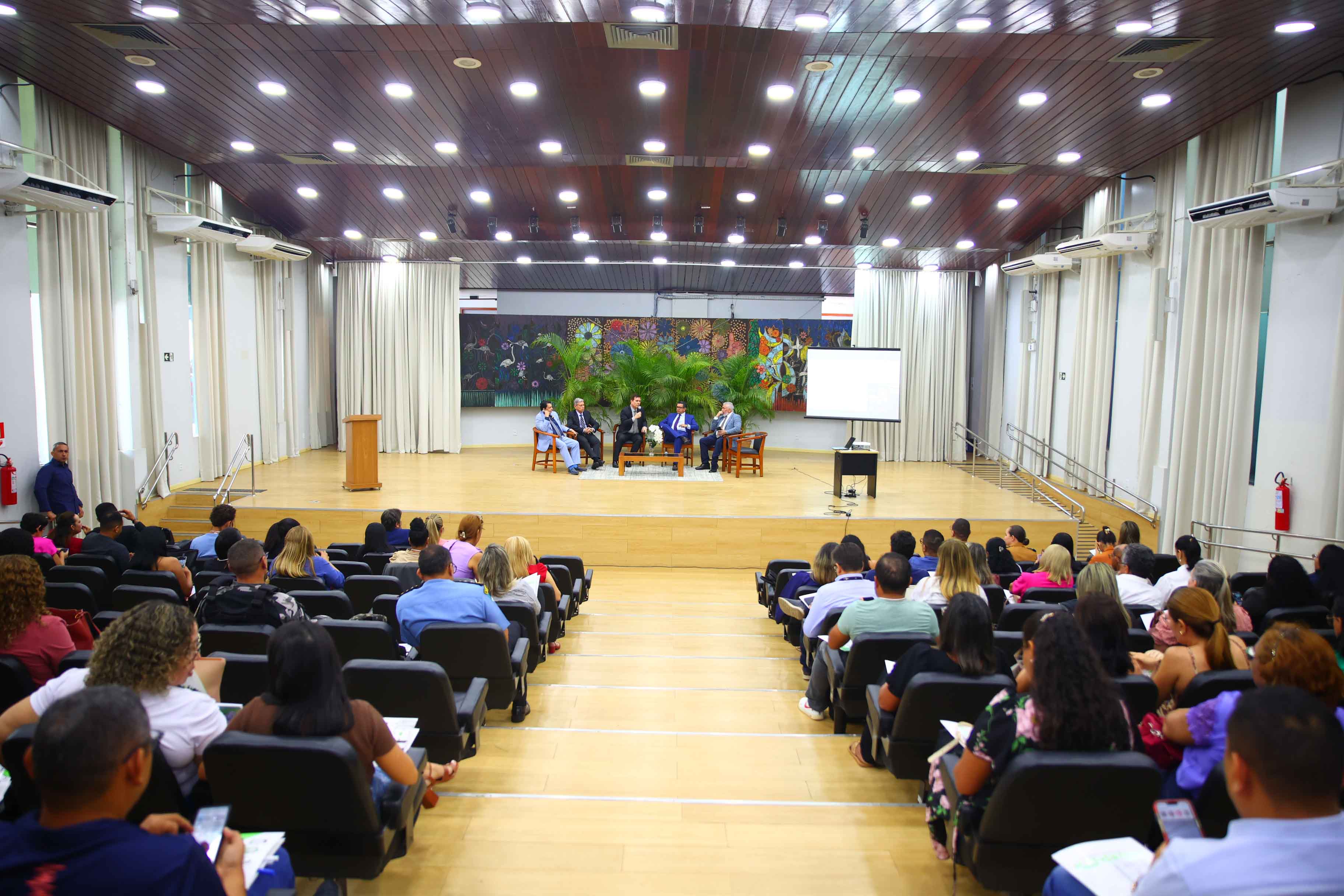 Imagem colorida mostra um painel de debate no palco de um auditório, onde cinco homens estão sentados em poltronas, de frente para a plateia. O auditório está cheio de pessoas sentadas em fileiras de cadeiras escuras, com um telão de projeção no canto direito do palco. Ao fundo, cortinas claras, um quadro abstrato e folhagens. Imagem colorida mostra um painel de debate no palco de um auditório, onde cinco homens estão sentados em poltronas, de frente para a plateia. O auditório está cheio de pessoas sentadas em fileiras de cadeiras escuras, com um telão de projeção no canto direito do palco. Ao fundo, cortinas claras, um quadro abstrato e folhagens.