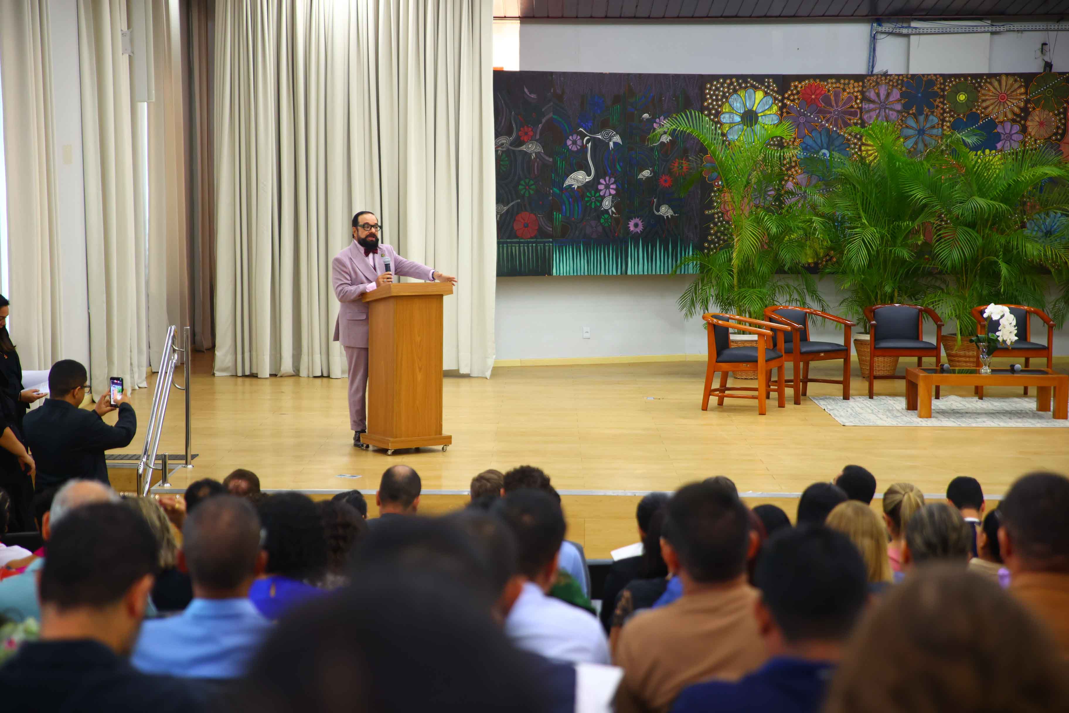 Imagem colorida mostra o presidente do TJRR, desembargador Leonardo Cupello, discursando em um púlpito de madeira, no palco de um auditório. Ele está no centro da foto, à frente de uma plateia lotada de pessoas sentadas. Ao fundo, cortinas claras, um quadro abstrato colorido com temas tropicais e um conjunto de poltronas e mesa de centro. Imagem colorida mostra o presidente do TJRR, desembargador Leonardo Cupello, discursando em um púlpito de madeira, no palco de um auditório. Ele está no centro da foto, à frente de uma plateia lotada de pessoas sentadas. Ao fundo, cortinas claras, um quadro abstrato colorido com temas tropicais e um conjunto de poltronas e mesa de centro.