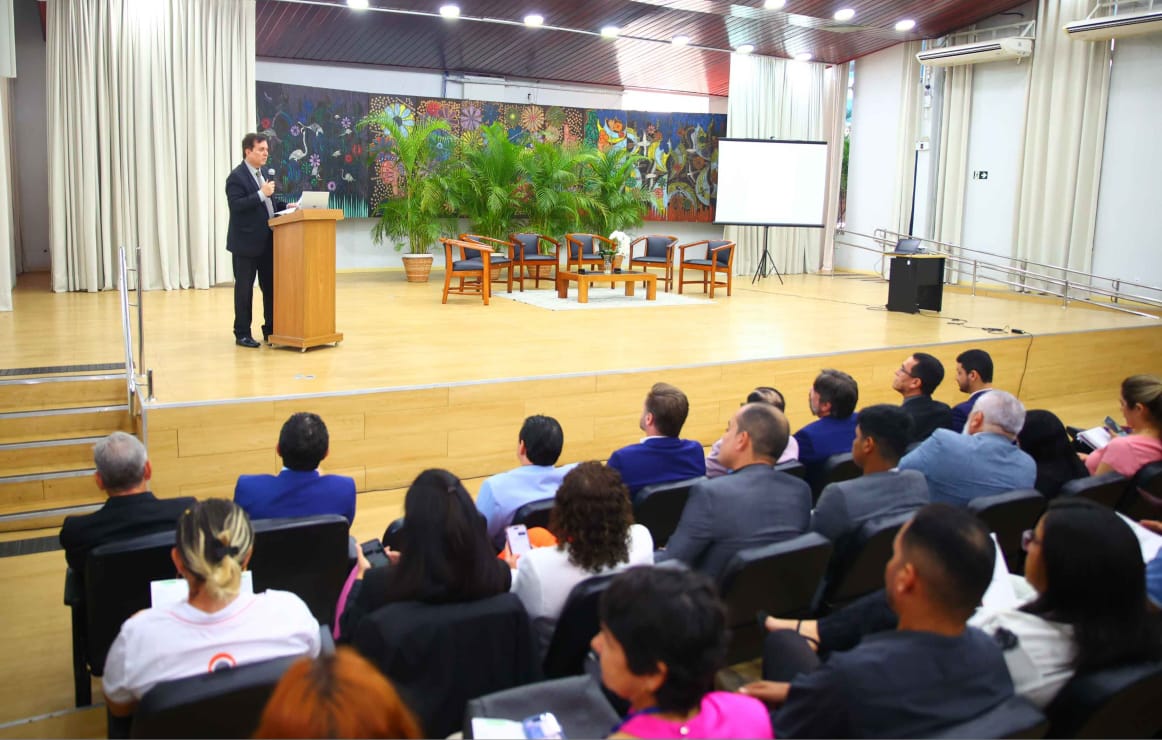 Imagem colorida mostra o juiz titular da Vepema, Alexandre Magno, discursando em um púlpito de madeira, no lado esquerdo do palco de um auditório. A plateia está sentada em cadeiras escuras, virada para o palco, e um telão de projeção está à direita. O fundo do palco tem uma decoração com cortinas claras, um quadro colorido e folhagens. Imagem colorida mostra o juiz titular da Vepema, Alexandre Magno, discursando em um púlpito de madeira, no lado esquerdo do palco de um auditório. A plateia está sentada em cadeiras escuras, virada para o palco, e um telão de projeção está à direita. O fundo do palco tem uma decoração com cortinas claras, um quadro colorido e folhagens.