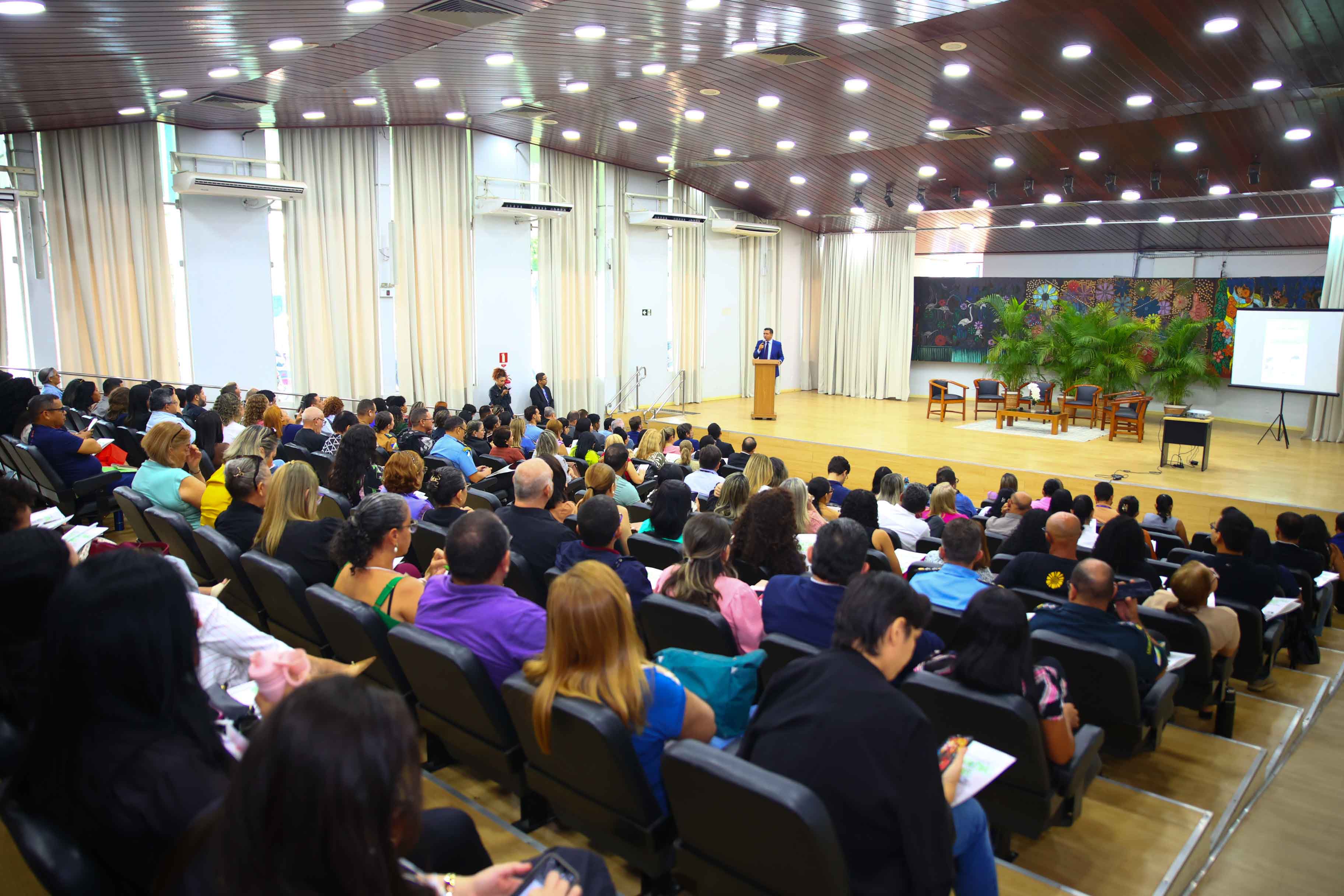 Imagem colorida mostra uma visão geral de um auditório, com um palestrante discursando em um púlpito no palco. A grande plateia ocupa as fileiras de cadeiras, sendo a maioria vista de costas, evidenciando o grande público do evento. A iluminação superior e o palco com cortinas e um painel de arte abstrata ao fundo são visíveis. Imagem colorida mostra uma visão geral de um auditório, com um palestrante discursando em um púlpito no palco. A grande plateia ocupa as fileiras de cadeiras, sendo a maioria vista de costas, evidenciando o grande público do evento. A iluminação superior e o palco com cortinas e um painel de arte abstrata ao fundo são visíveis.