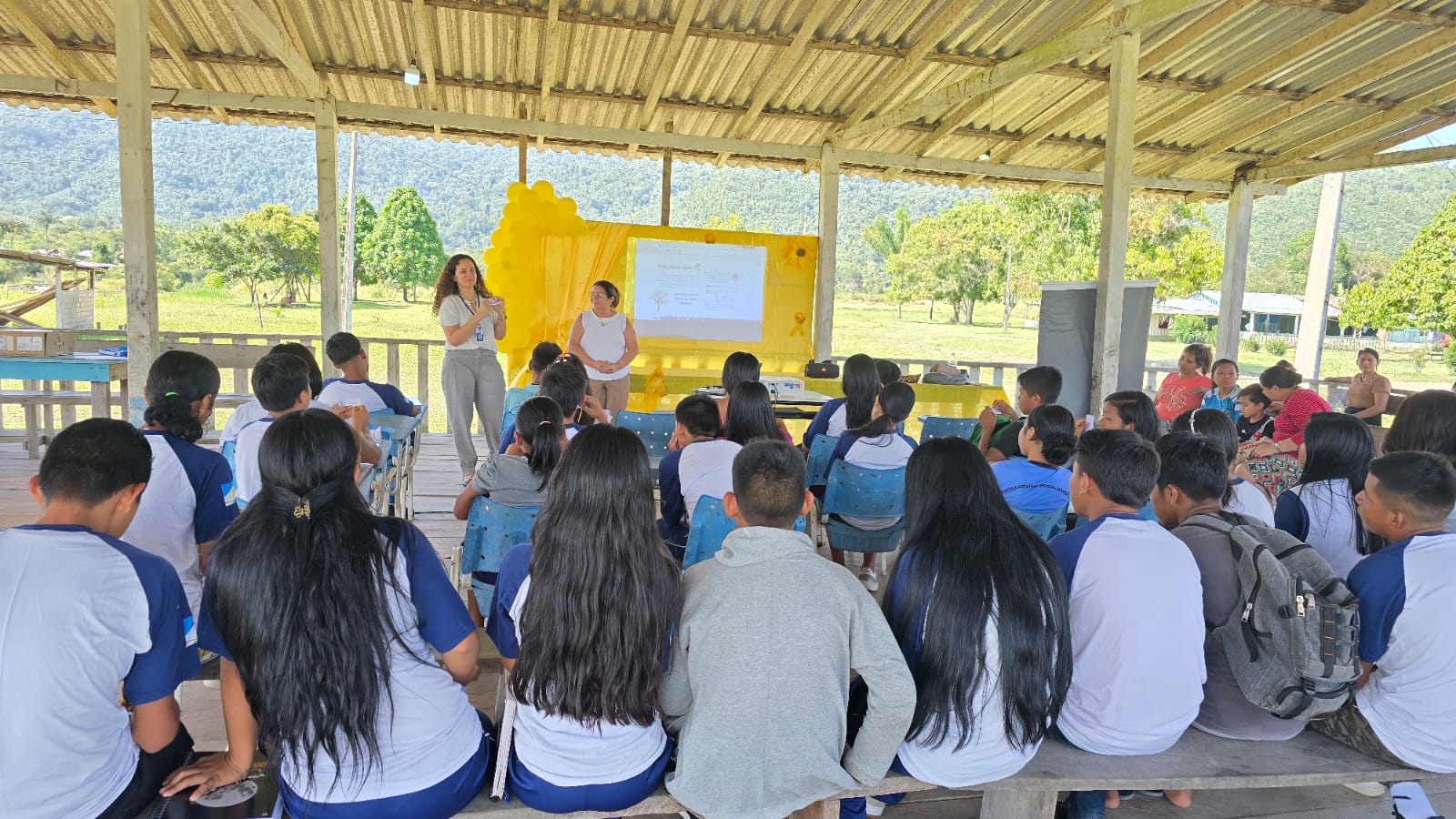 A imagem mostra um grupo de estudantes assistindo a uma apresentação em um espaço coberto de madeira, com laterais abertas. Eles estão sentados em cadeiras azuis e também em bancos de madeira. À frente, duas mulheres estão em pé conduzindo a atividade, uma delas segurando um microfone. Atrás delas há uma tela de projeção com slides e uma decoração amarela com balões. Ao fundo, pela estrutura aberta, é possível ver árvores, gramado e montanhas sob um céu claro.