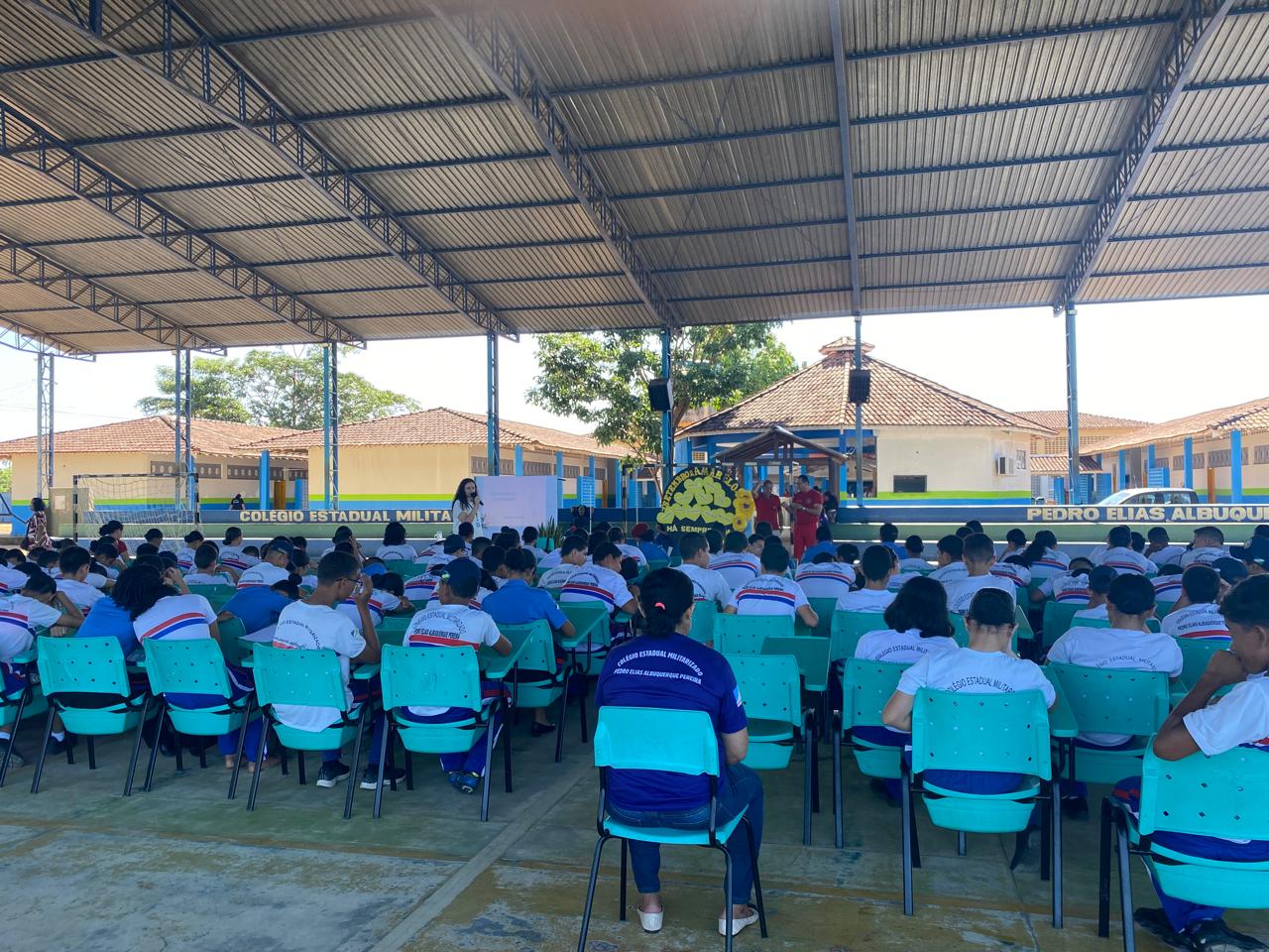 A imagem mostra um grande grupo de estudantes sentados em cadeiras azul-turquesa, sob uma quadra coberta. Eles vestem uniformes brancos e azuis do Colégio Estadual Militar Pedro Elias de Albuquerque. À frente, há um pequeno palco com faixas verdes e azuis, onde algumas pessoas, incluindo uma mulher de roupa clara e integrantes vestidos de vermelho, conduzem uma atividade. Ao fundo, é possível ver os prédios escolares de telhados marrons e paredes amarelas, cercados por árvores e céu azul.