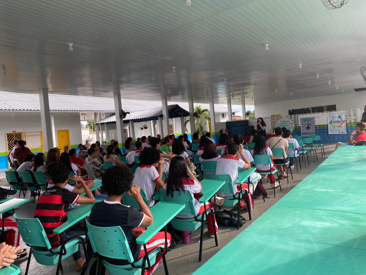 A imagem mostra um grupo de estudantes e adultos participando de uma atividade em um espaço coberto e bem iluminado. As pessoas estão sentadas em cadeiras e carteiras verde-água, voltadas para a frente, onde uma mulher fala ao público com o auxílio de um microfone. Atrás dela, há banners com logotipos e informações do Programa Justiça Comunitária fixados na parede. Os participantes vestem uniformes escolares em branco, vermelho e preto. O ambiente é amplo, com piso de concreto e estrutura moderna em tons de branco, azul e verde.