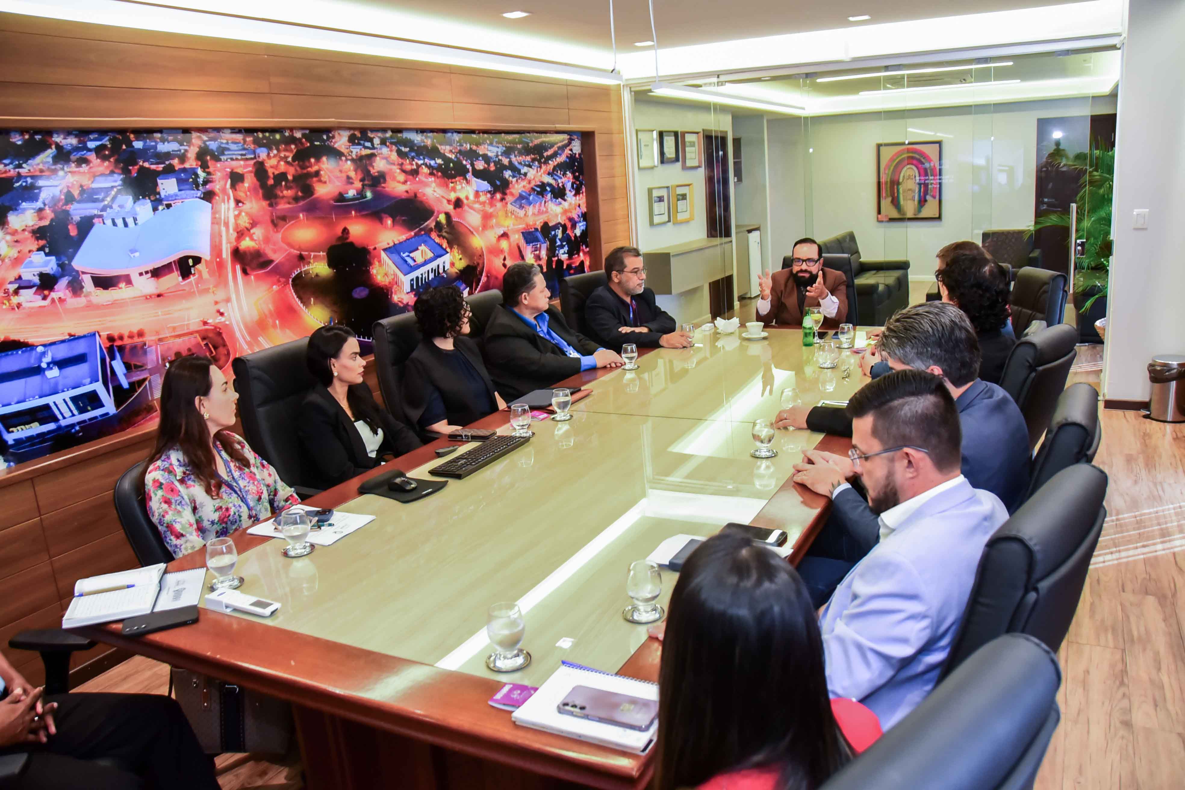 Imagem mostra uma reunião em uma sala ampla e bem iluminada, com uma grande mesa de madeira e tampo de vidro. Cerca de doze pessoas estão sentadas ao redor. No centro da mesa, há copos com água, notebooks, anotações e um teclado. As pessoas vestem trajes formais, como ternos, blazers e camisas sociais. Ao fundo, há uma parede de vidro com persianas verticais e uma planta decorativa. O ambiente tem um ar institucional e profissional, sugerindo uma reunião de trabalho ou encontro entre gestores. Imagem mostra uma reunião em uma sala ampla e bem iluminada, com uma grande mesa de madeira e tampo de vidro. Cerca de doze pessoas estão sentadas ao redor. No centro da mesa, há copos com água, notebooks, anotações e um teclado. As pessoas vestem trajes formais, como ternos, blazers e camisas sociais. Ao fundo, há uma parede de vidro com persianas verticais e uma planta decorativa. O ambiente tem um ar institucional e profissional, sugerindo uma reunião de trabalho ou encontro entre gestores.