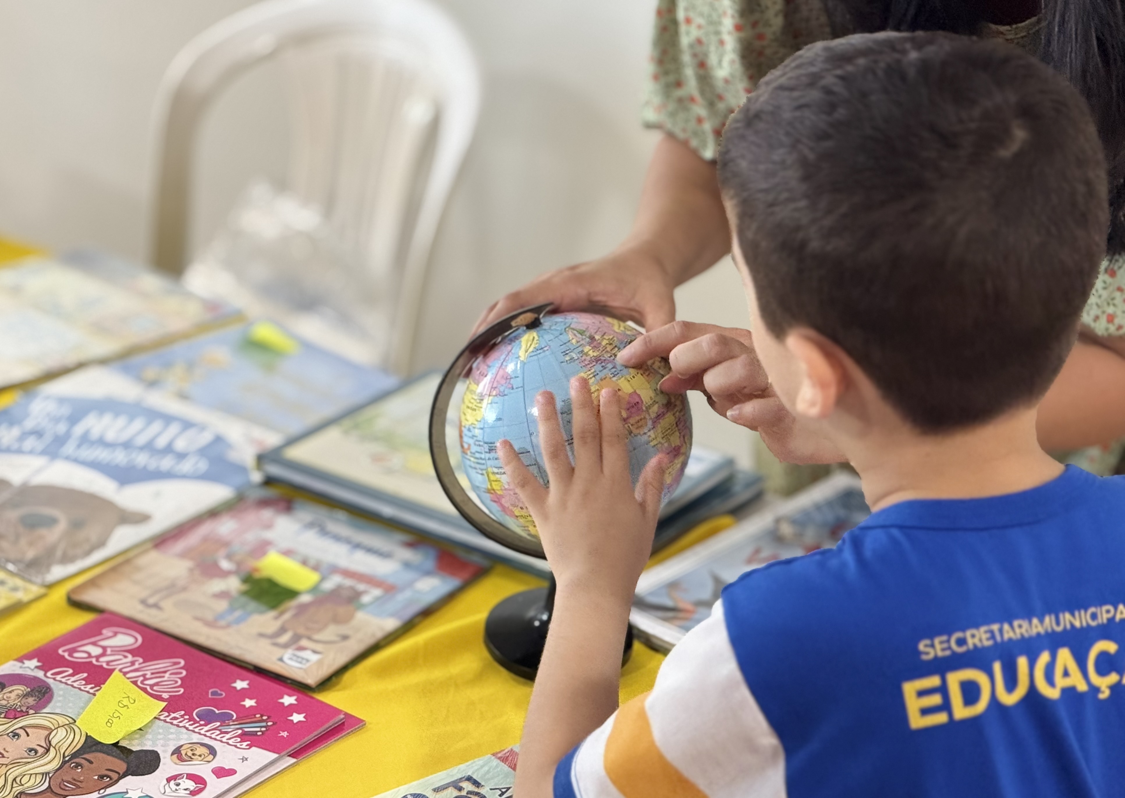 criança em atividade educativa, apontando para um globo terrestre sobre uma mesa com livros, em uma clara referência à aprendizagem e acesso à cultura.