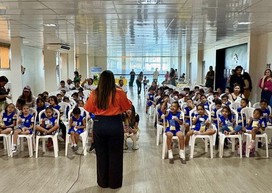 Crianças uniformizadas assistindo atentas a apresentações em um salão. Uma cena mostra uma palestrante interagindo com a plateia; a outra, uma performance de um casal (um vestido de cozinheiro) no palco decorado com o logo do TJRR/EJURR.