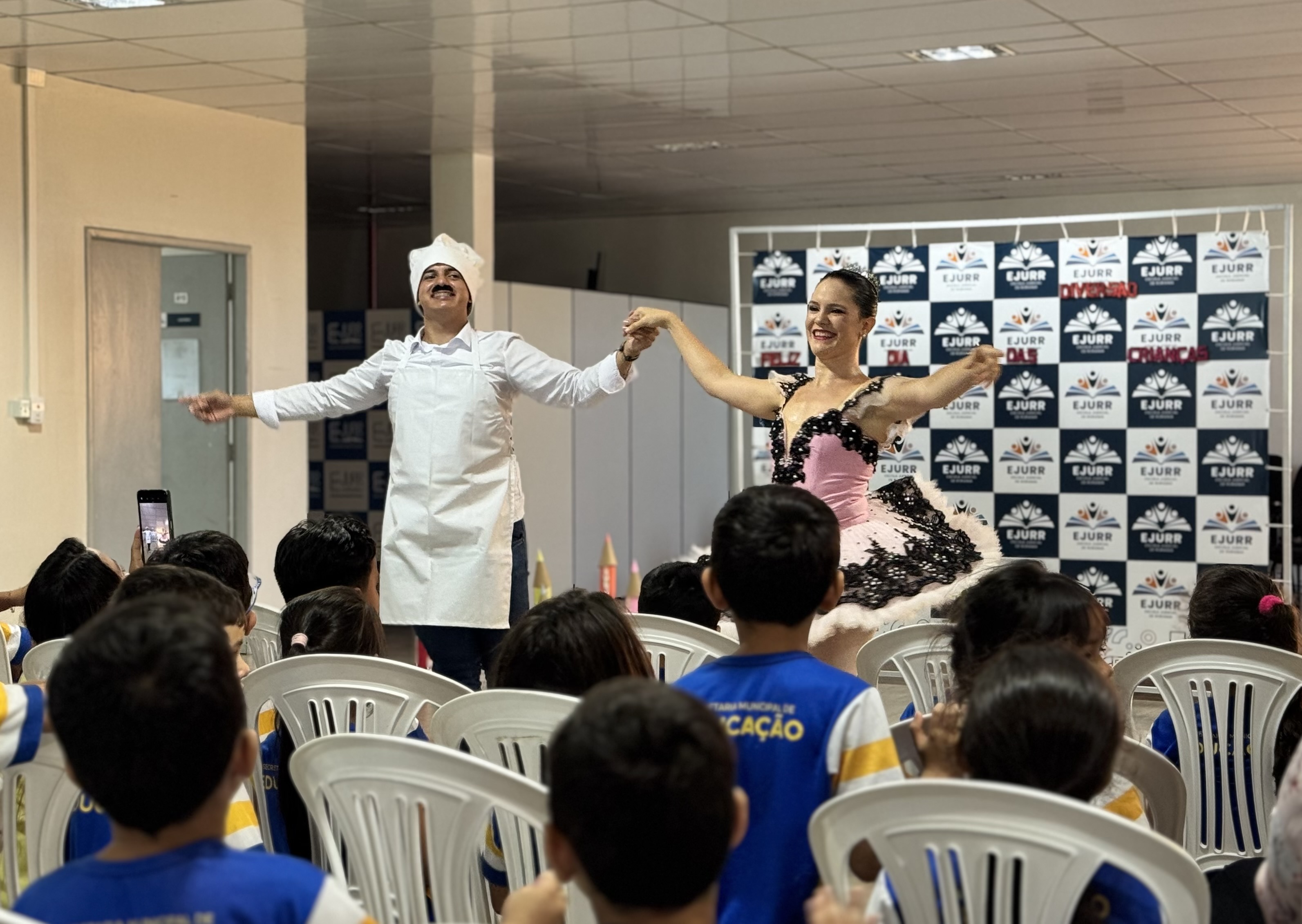 Crianças assistem a uma apresentação de dança ou teatro, onde um homem vestido de cozinheiro e uma mulher com vestido de festa se apresentam em frente a um painel temático do TJRR/EJURR.