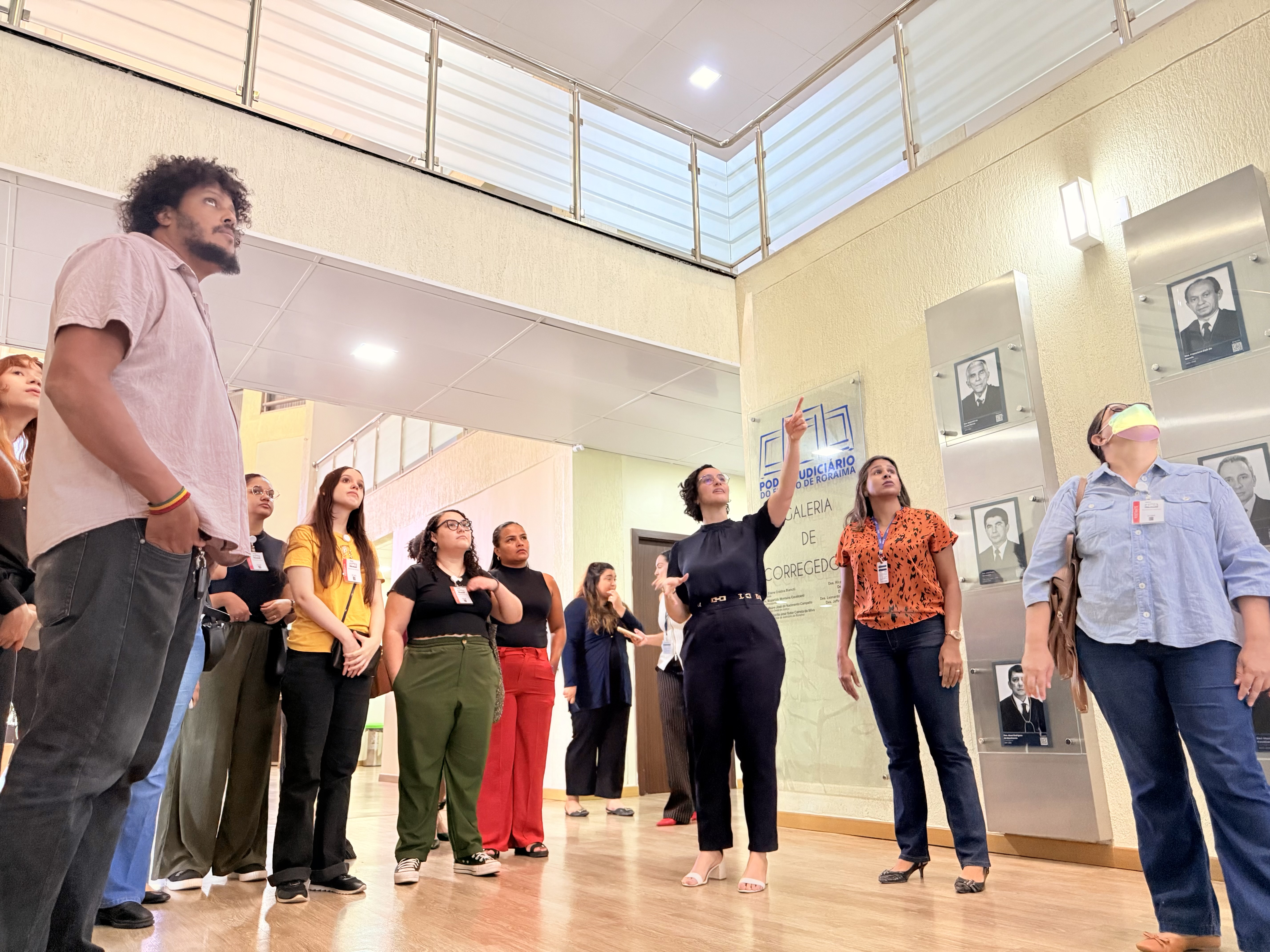 Imagem colorida mostra um grupo de pessoas em pé em um corredor claro com piso de madeira, olhando para cima. No centro, uma mulher de cabelo cacheado em um vestido escuro gesticula enquanto fala. Há quadros com fotos de pessoas na parede à direita.