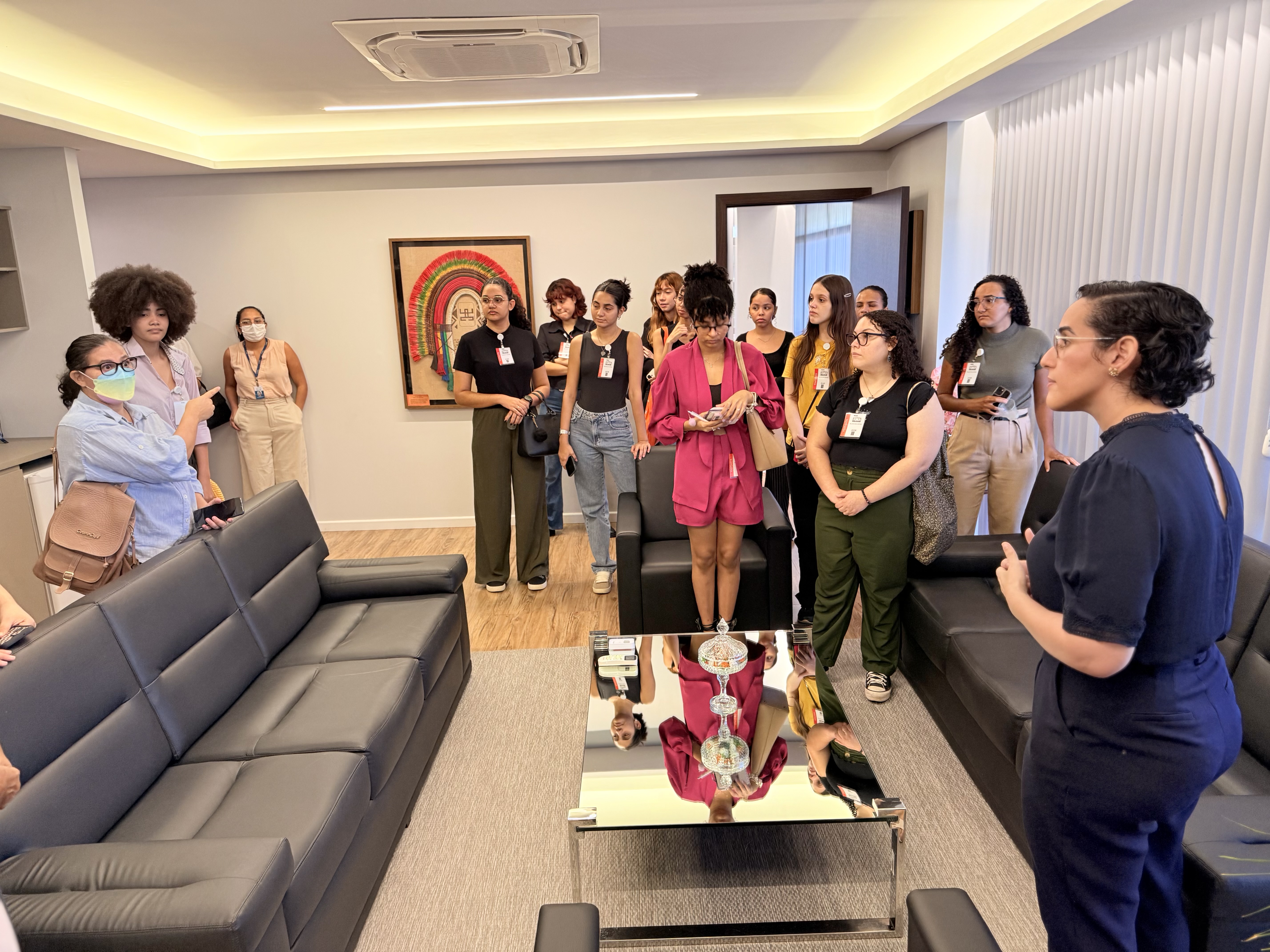 Imagem colorida mostra um grupo de pessoas em uma sala de estar moderna, com sofás pretos. Uma mulher de cabelo cacheado em um vestido escuro gesticula enquanto fala para o grupo que está à sua frente. Há um quadro com arte indígena na parede ao fundo.